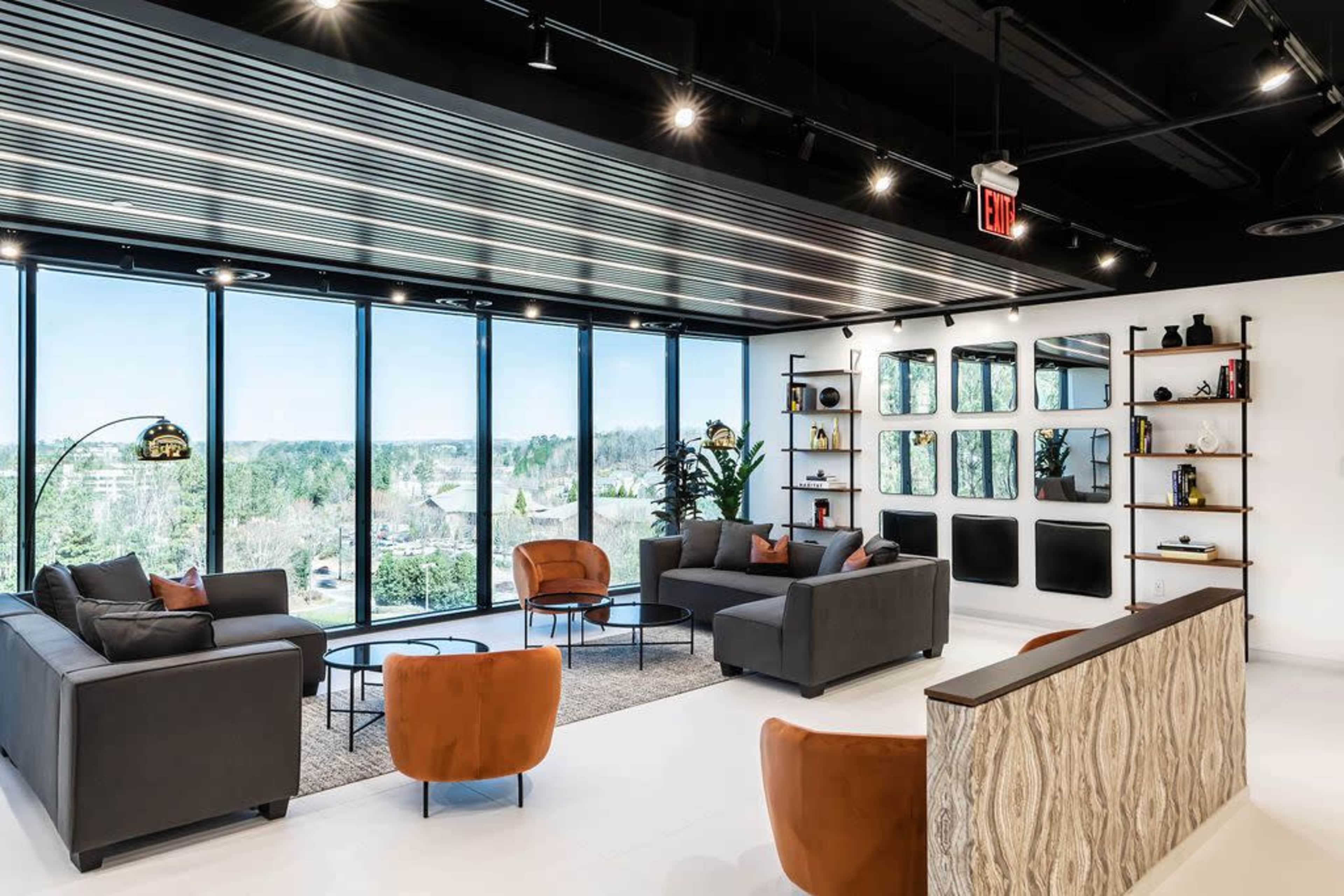 The image shows a modern office lounge with two dark sofas, orange accent chairs, a glass coffee table, and large windows offering a view of the landscape outside.