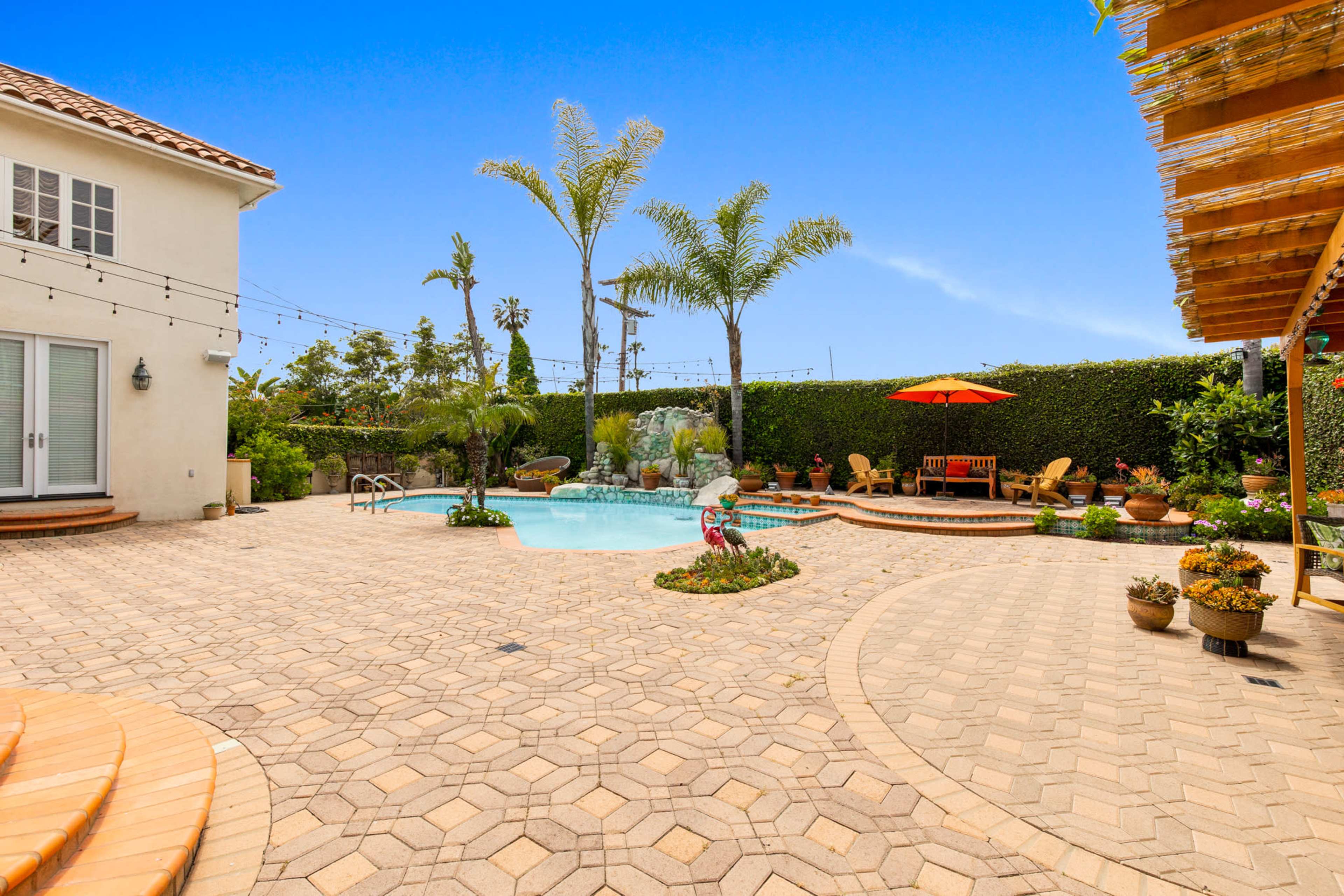A pool area surrounded by tropical plants, lounge chairs, and potted flowers on a paved patio.