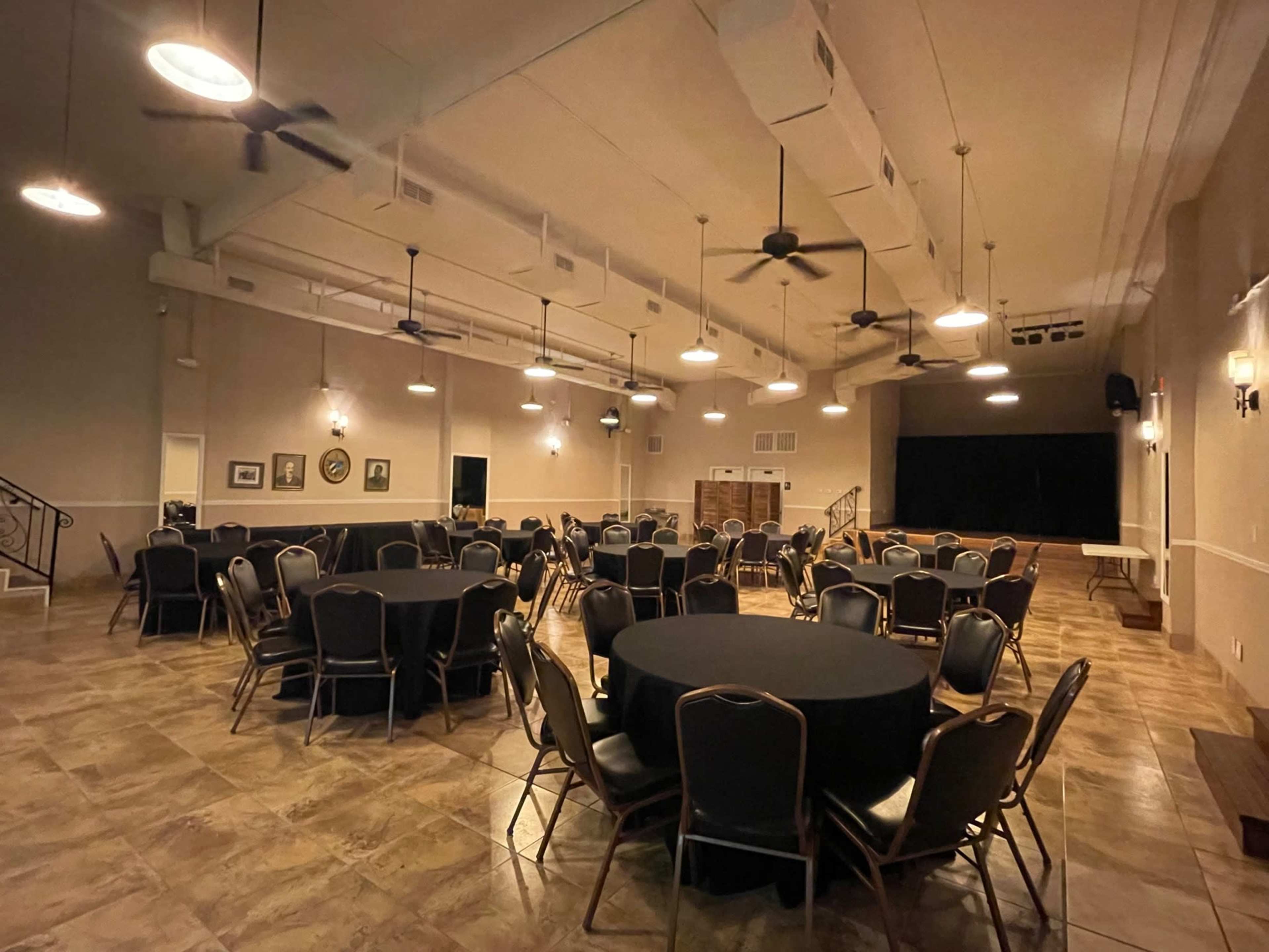 The interior of a large event space features multiple round tables with black tablecloths arranged on a tiled floor under ceiling fans and industrial lighting.