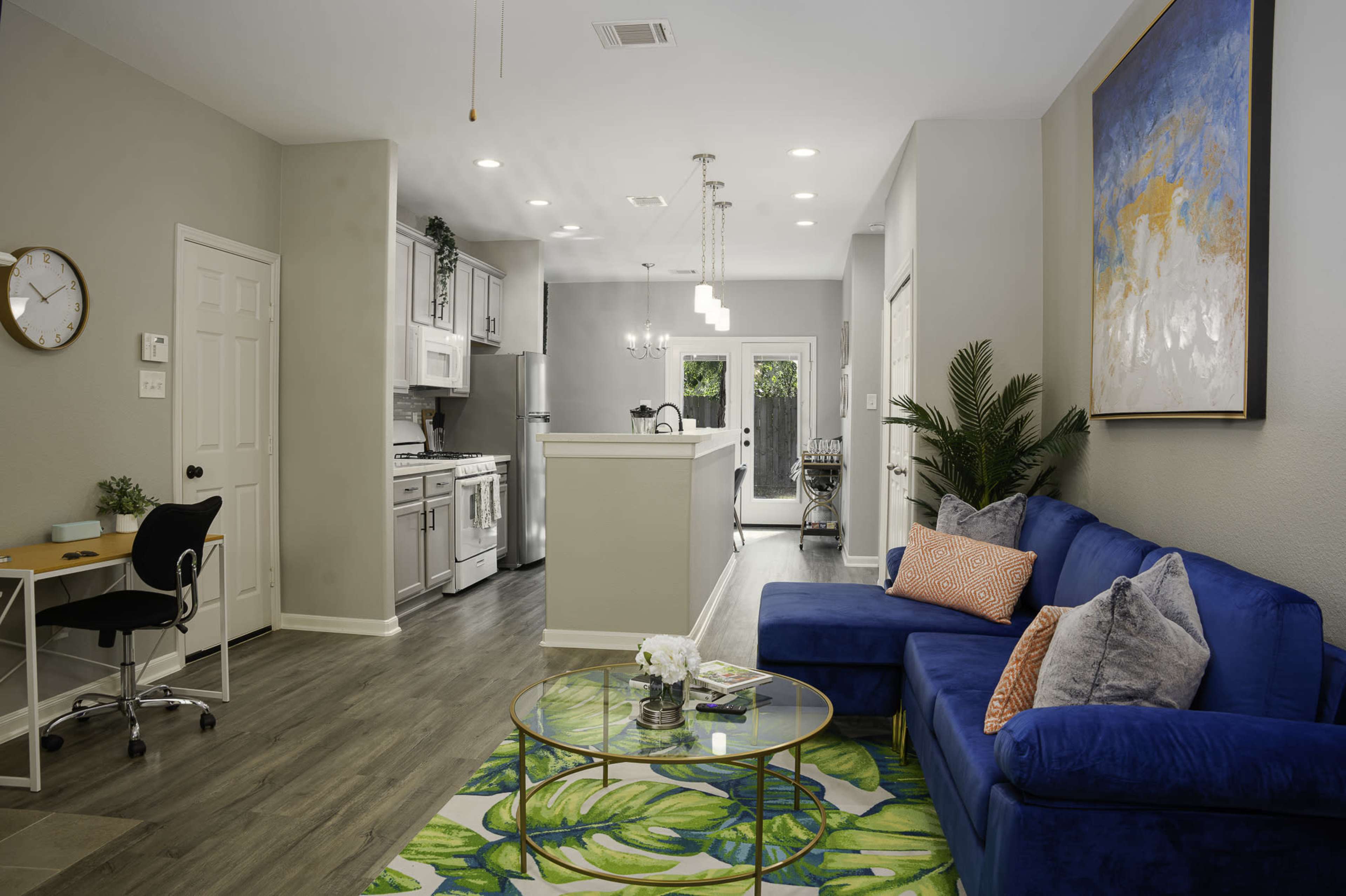 A modern living room features a blue couch, a glass coffee table, and a small workspace, with an open kitchen visible in the background.