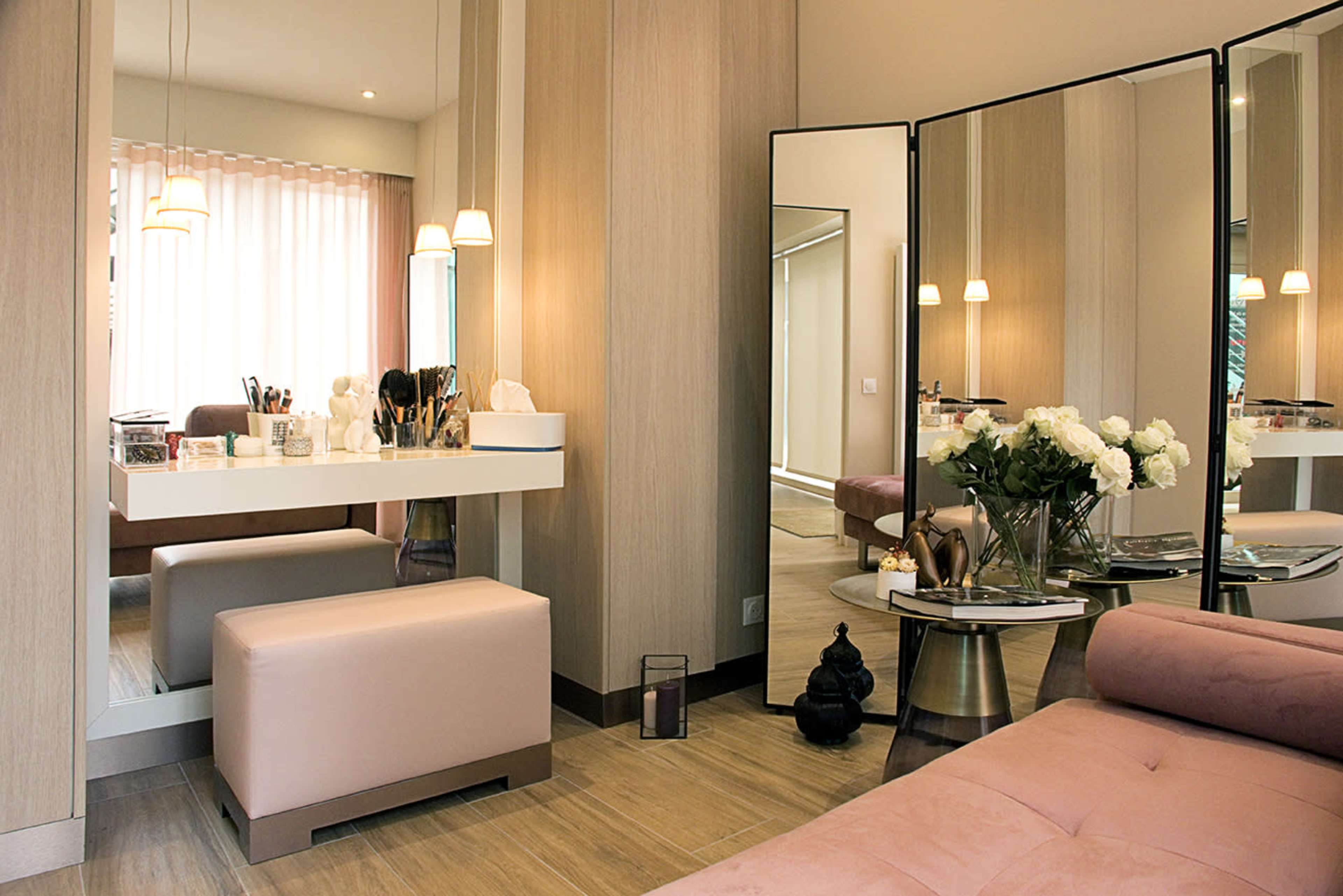 A well-decorated room featuring a makeup vanity with brushes and cosmetics, a plush pink ottoman, and a mirrored partition reflecting floral arrangements and soft lighting.