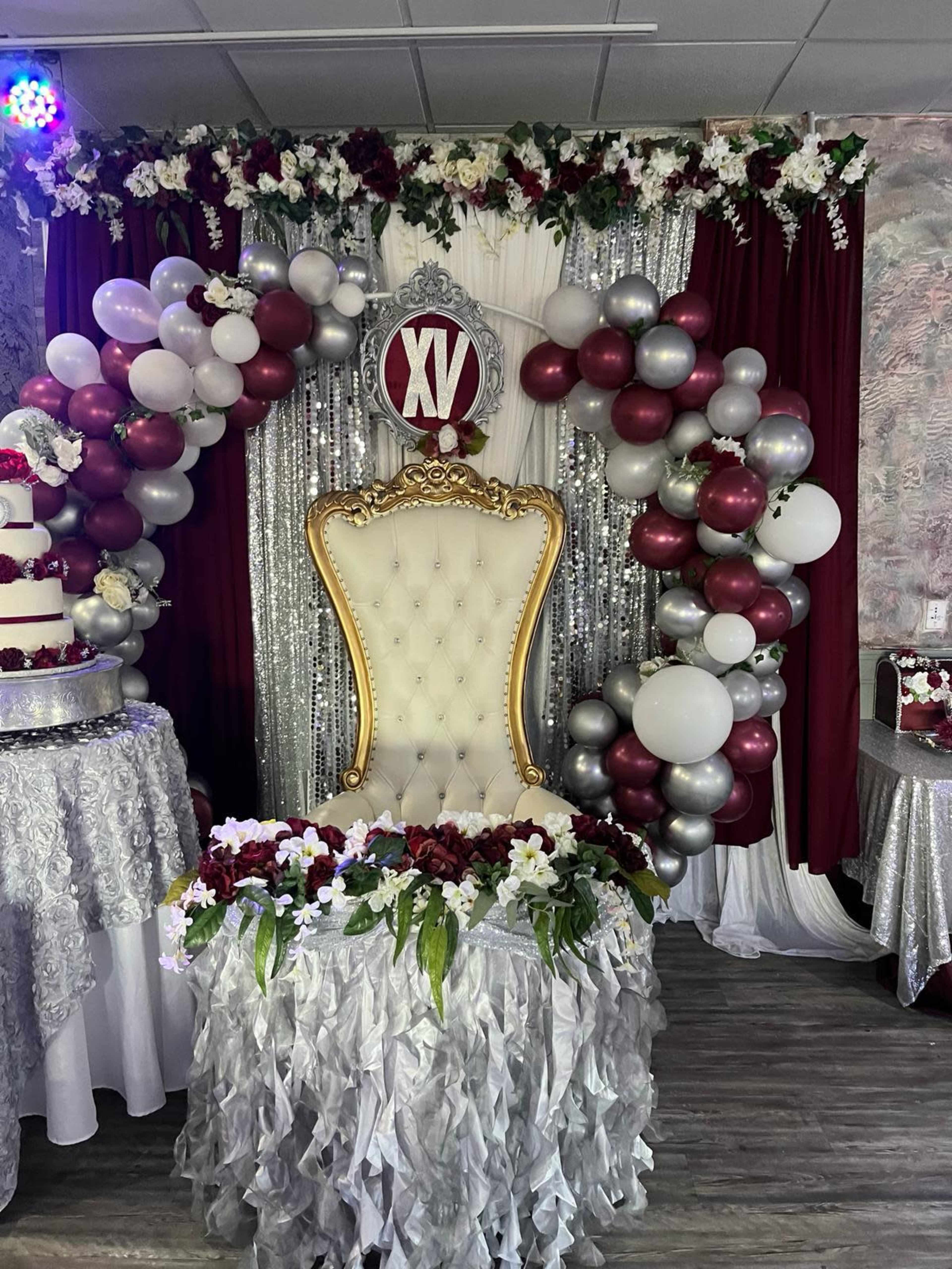 A decorated event space with a lavish throne chair surrounded by a floral and balloon arch, complete with a "XV" sign above it.