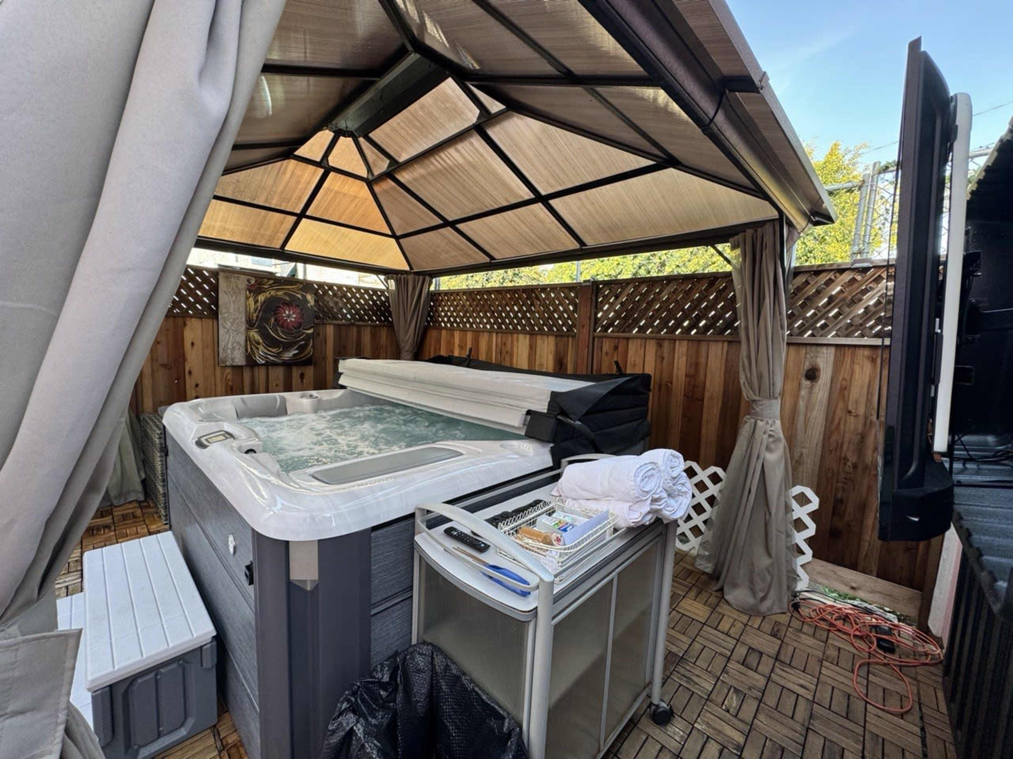 A hot tub covered by a canopy, surrounded by wooden fencing and equipped with a towel rack and a table with supplies.