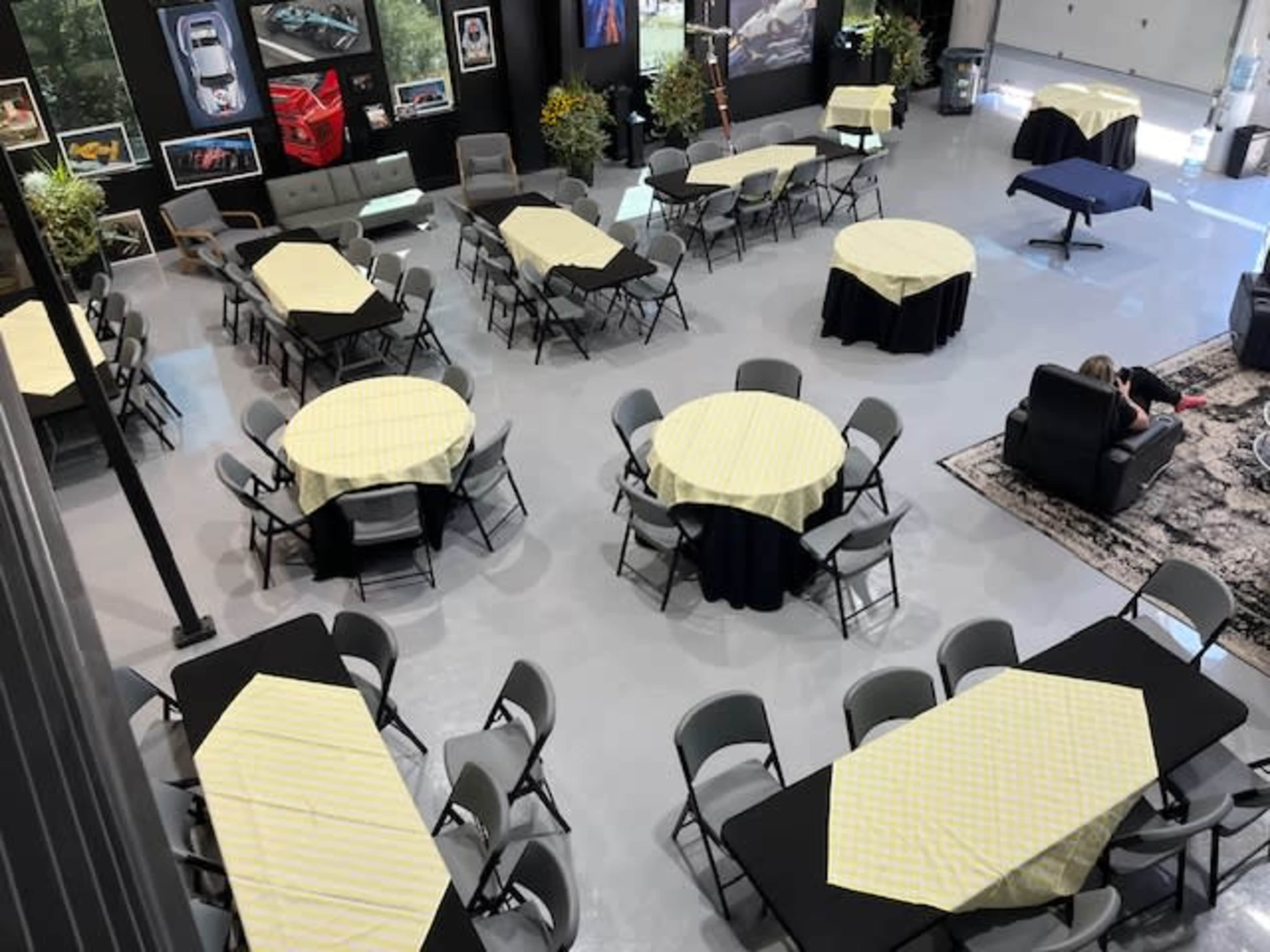 A spacious room features several round tables covered with yellow checkered tablecloths and black cloths, arranged around a seating area with chairs and a couch.