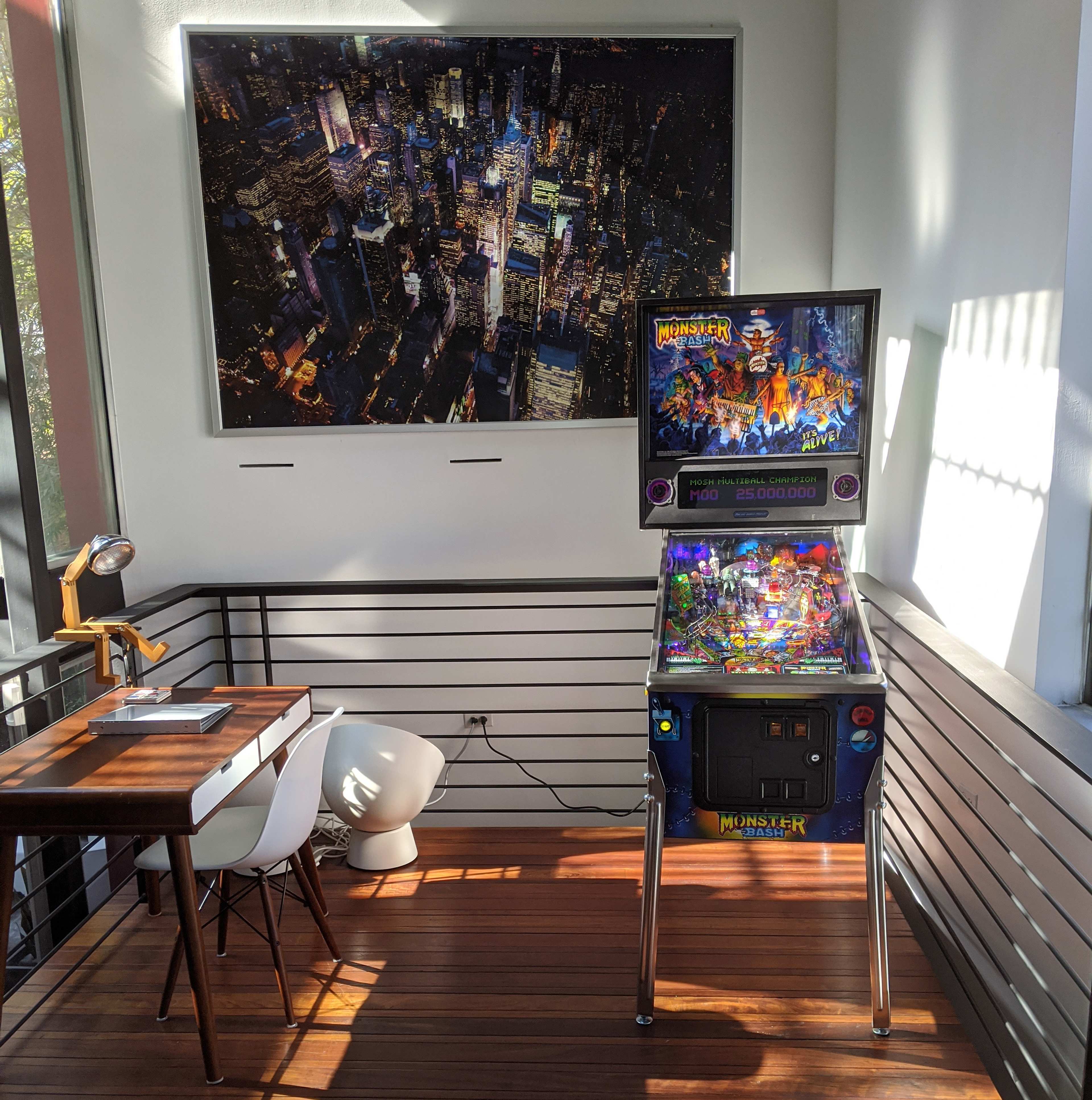 A pinball machine titled "Monster" is positioned next to a modern desk and a white chair in a sunlit corner of a room featuring a large cityscape photograph on the wall.
