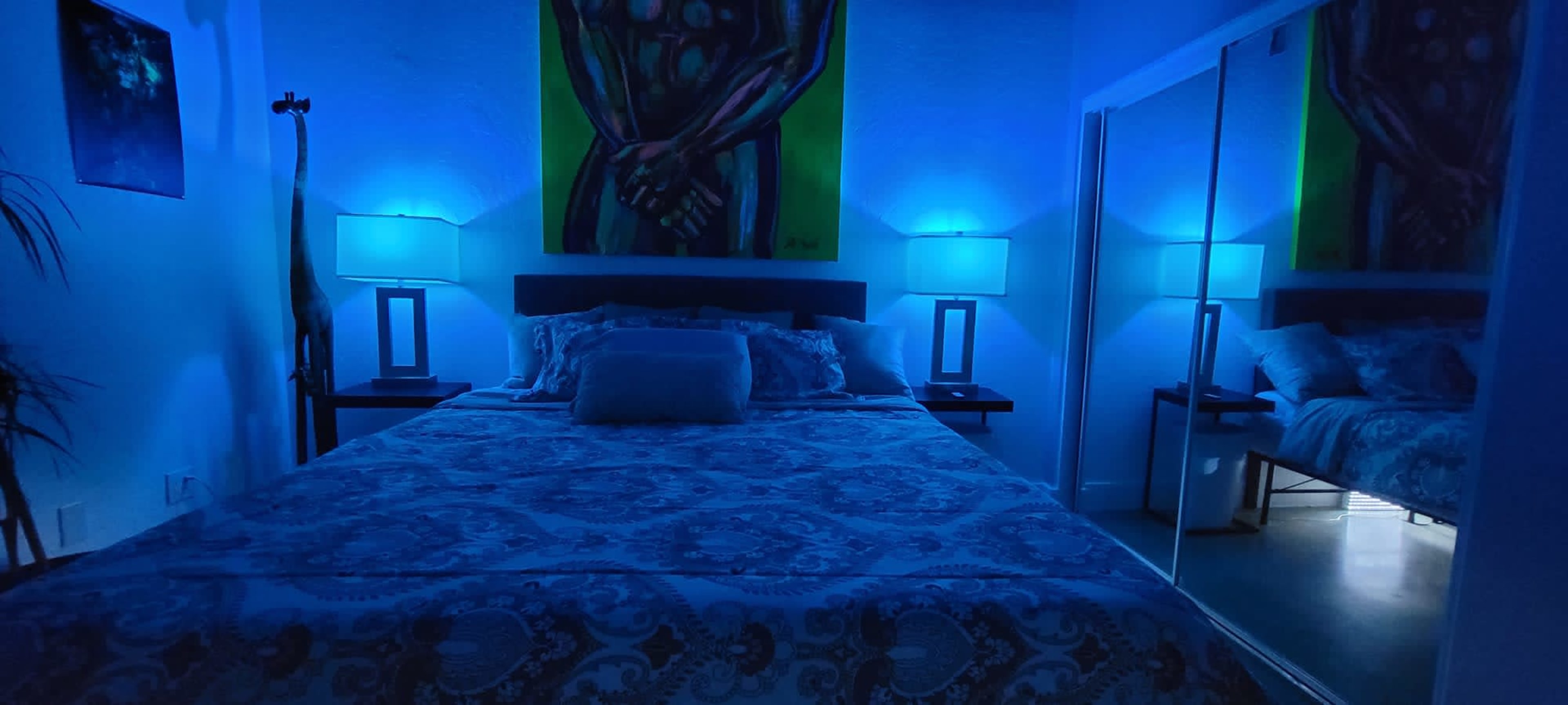 A bedroom features a bed with a patterned coverlet, illuminated by blue lights from two bedside lamps, and a large painting on the wall.
