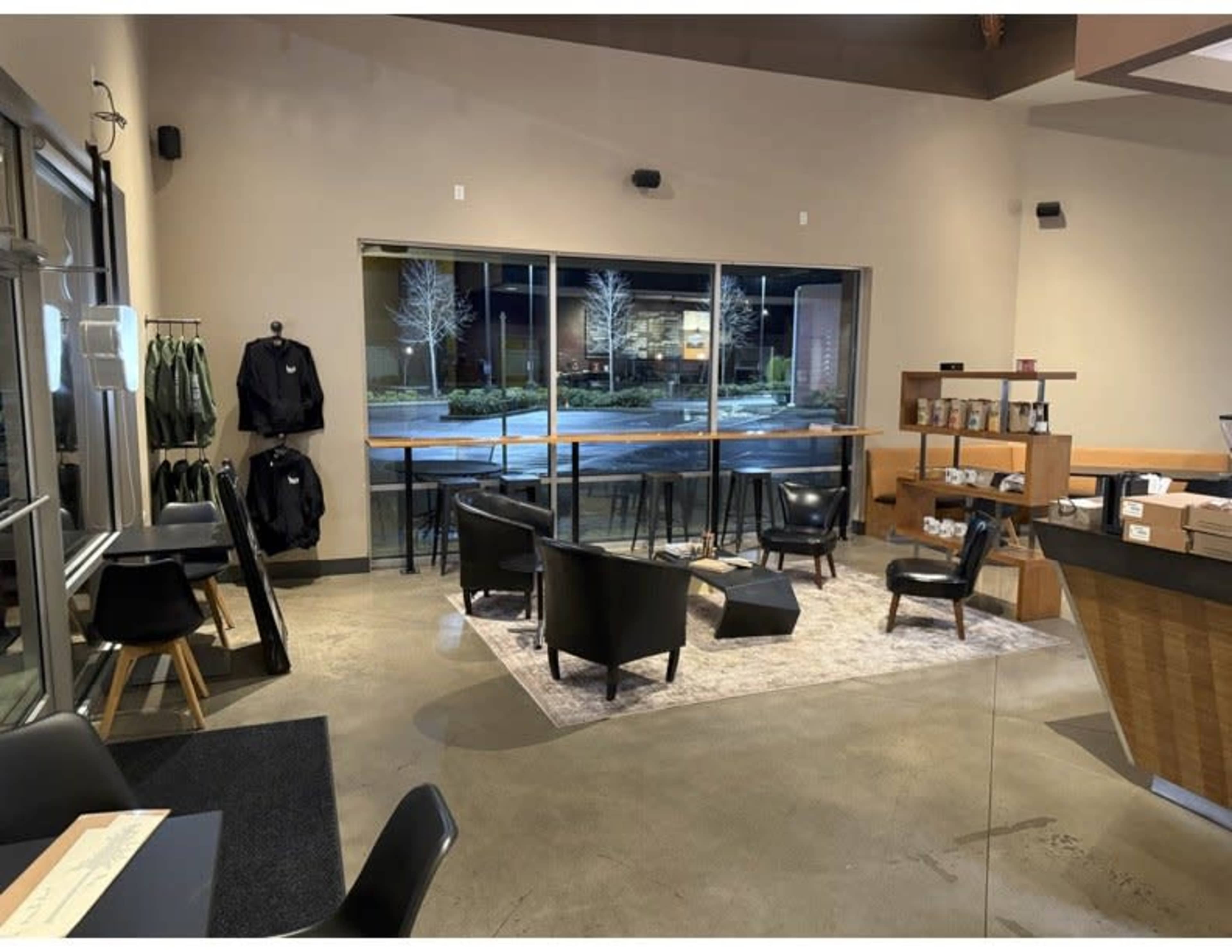 The image shows a modern indoor lounge area with black chairs and a small table, situated next to large windows facing an outdoor space.