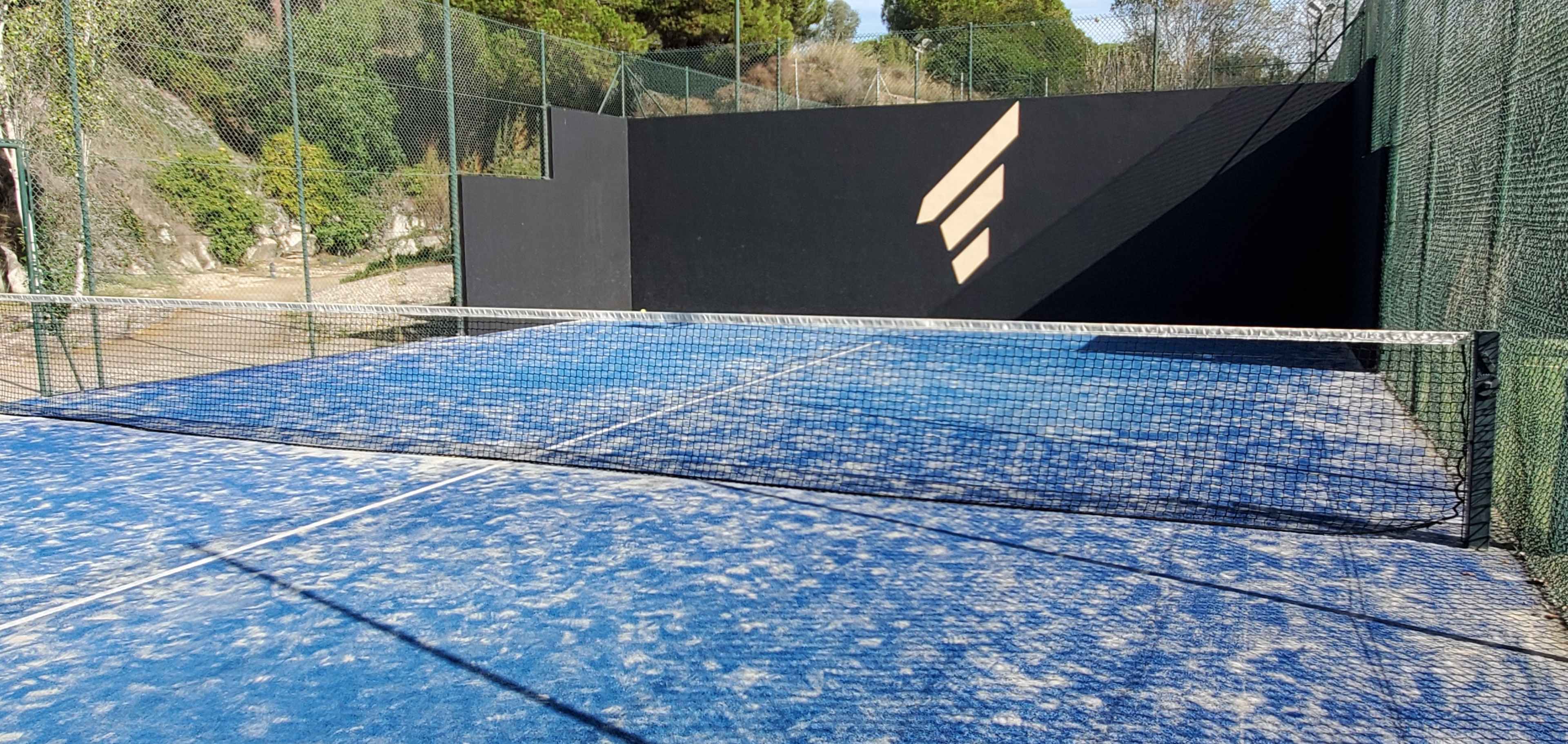 A blue tennis court enclosed by green fencing and a black wall, with sunlight creating diagonal shadows on the surface.