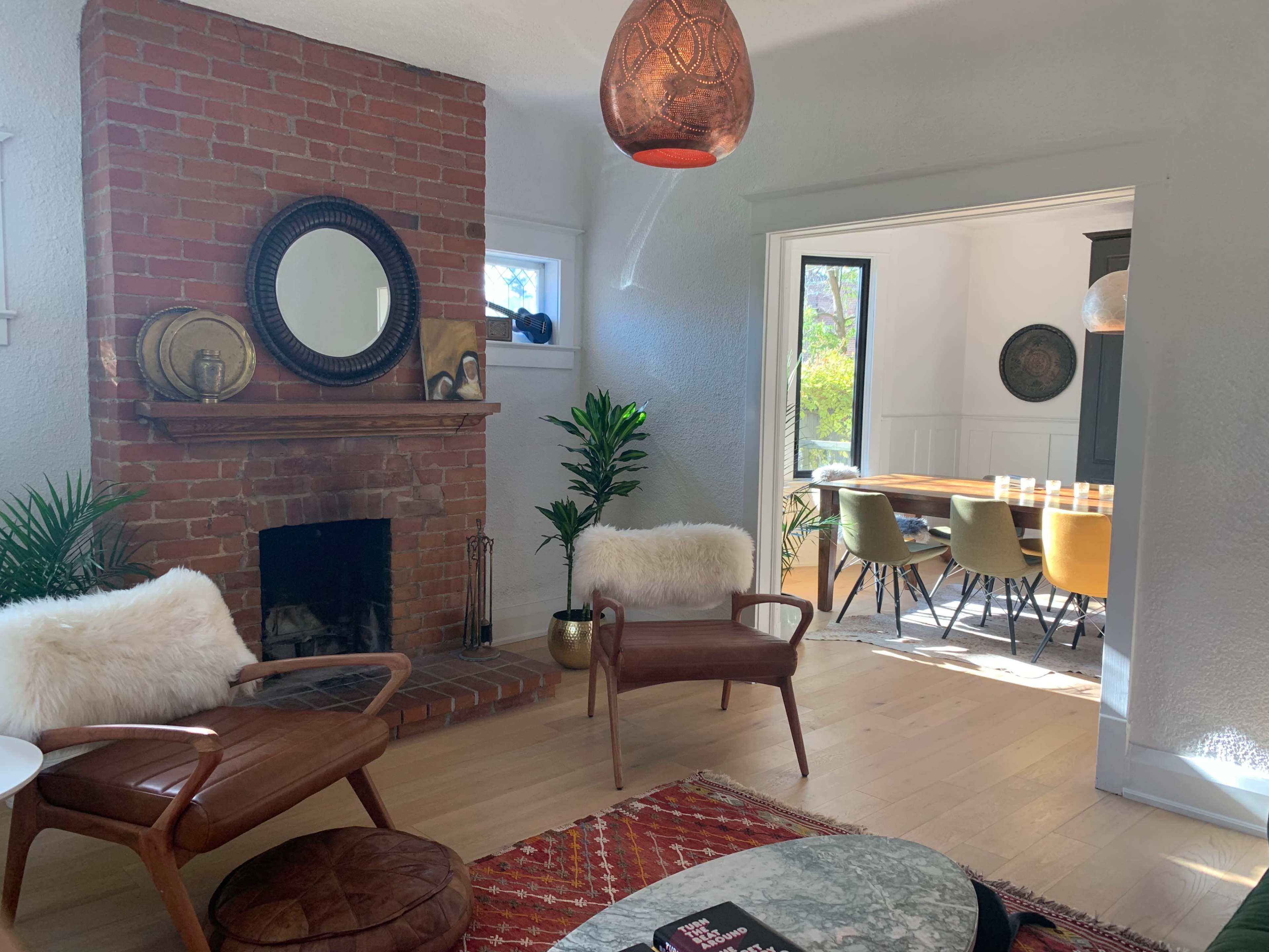 A cozy living room features a brick fireplace with a circular mirror above it, two mid-century modern chairs, and a view of a dining area with a large table and colorful chairs.