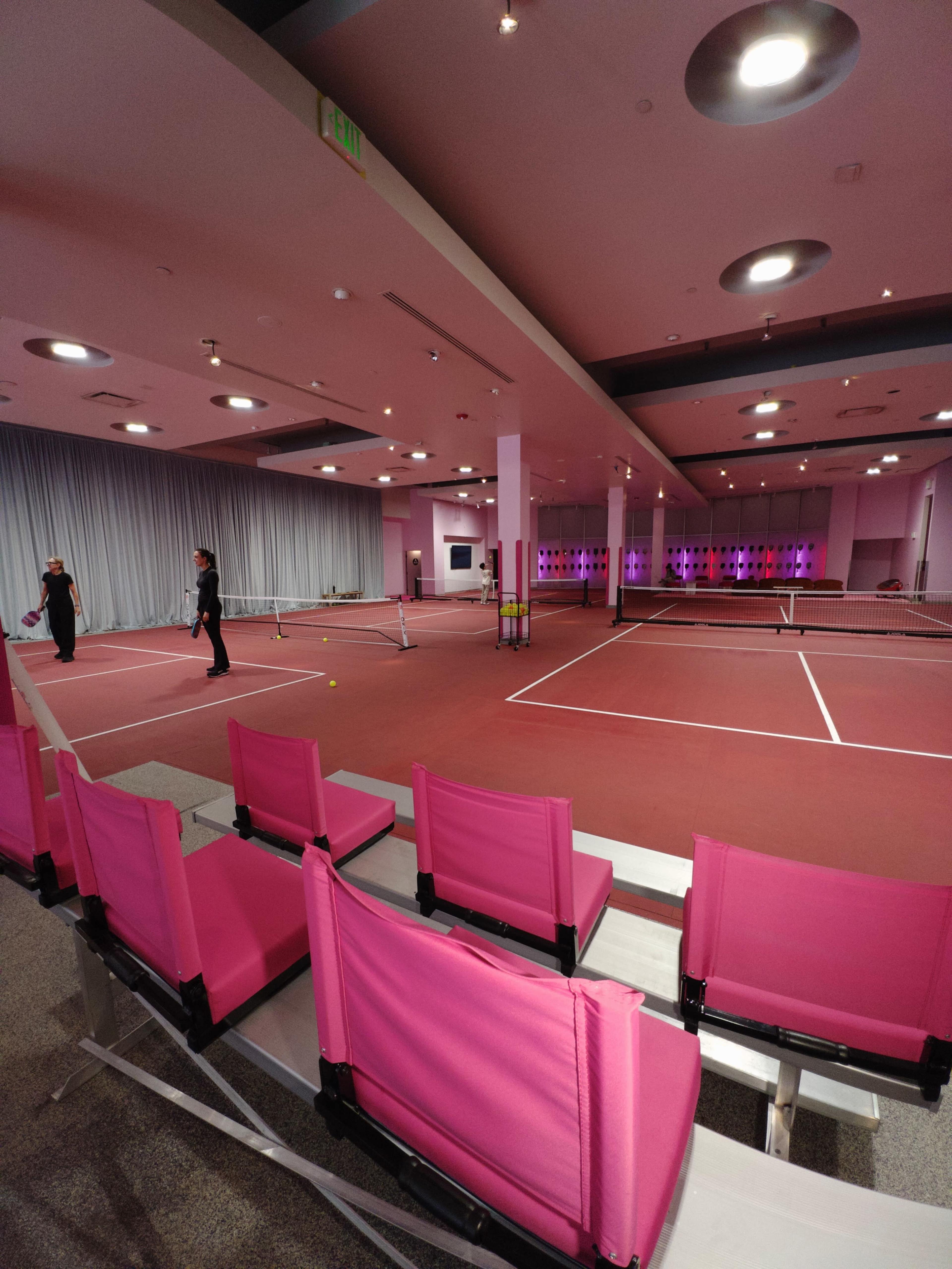 An indoor tennis court with pink seating, featuring two players practicing on the court.