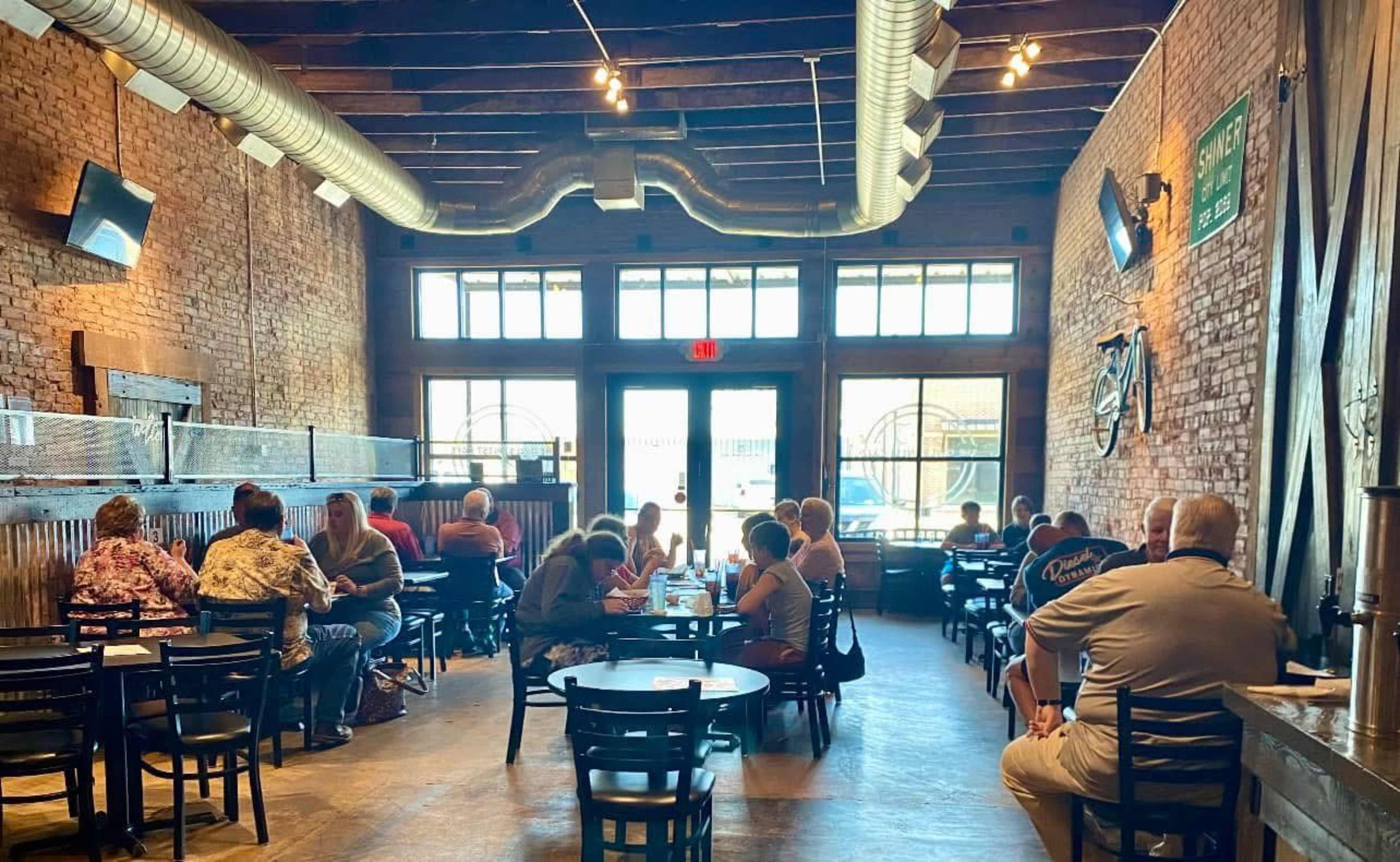 The image shows a restaurant interior with brick walls, wooden beams, and several tables occupied by diners.