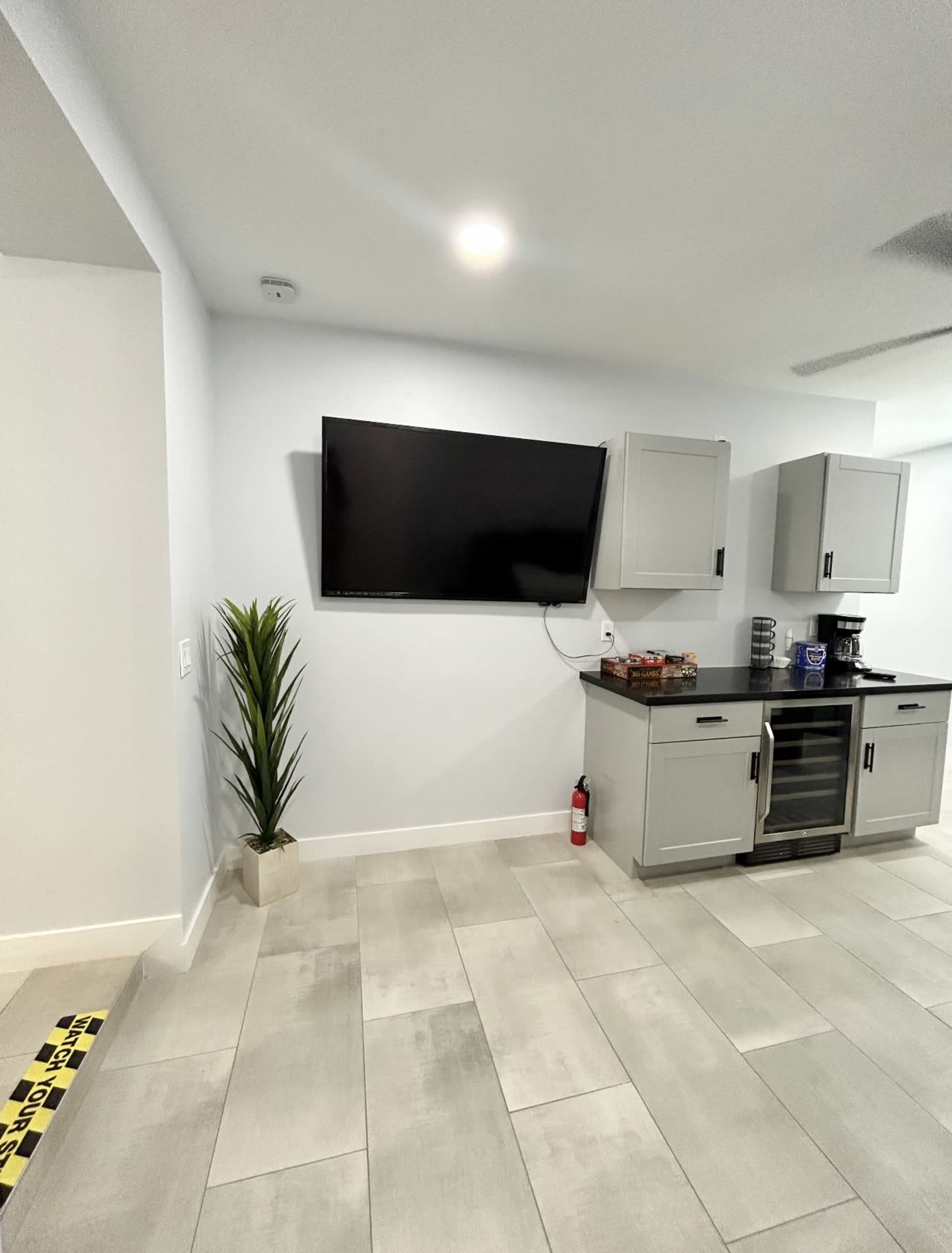 The image shows a modern, minimalist interior with a flat-screen TV mounted on a light-colored wall, accompanied by a small kitchenette and a decorative plant in the corner.