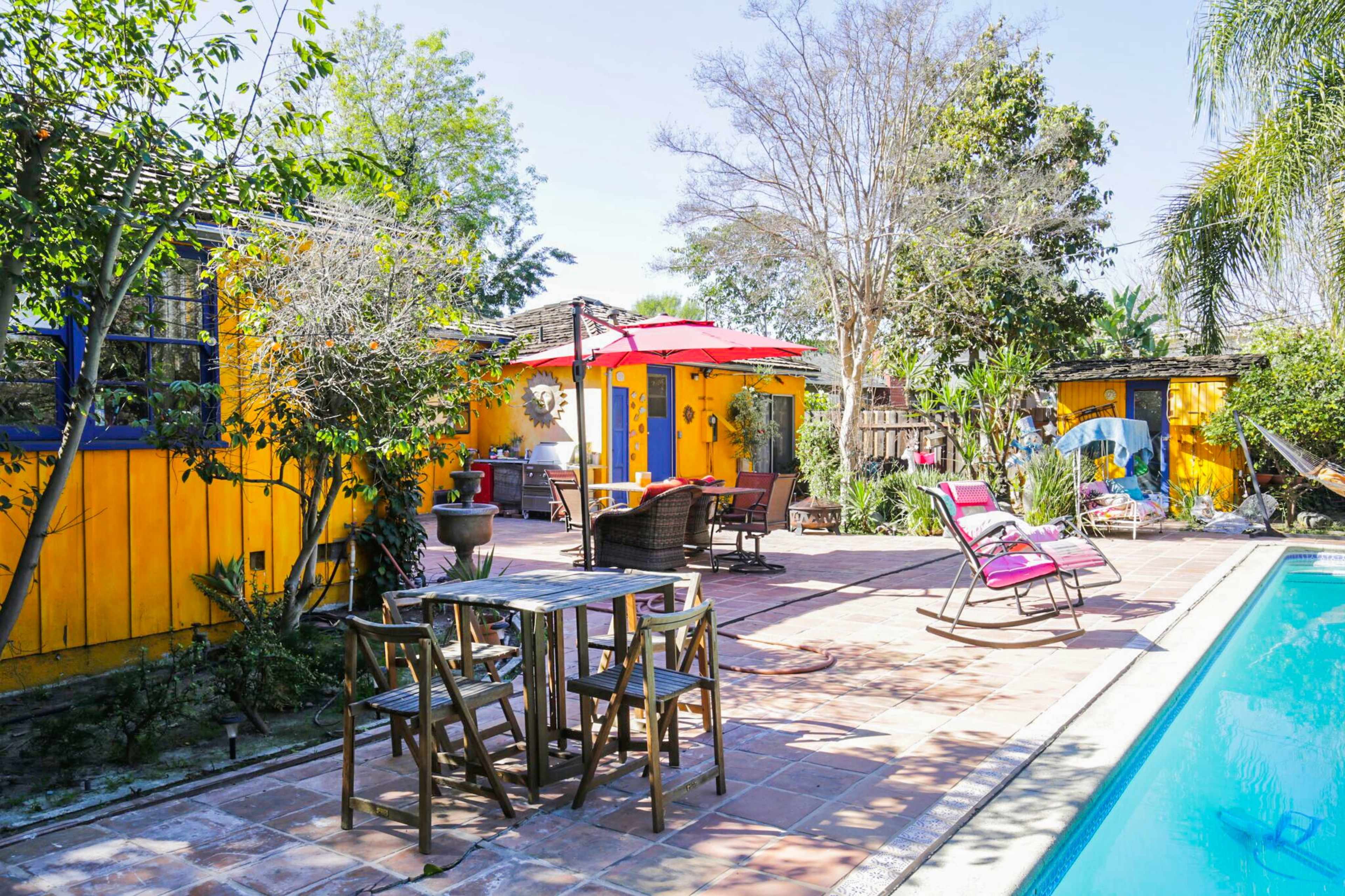A vibrant outdoor area features a swimming pool, a dining table with four chairs, and lounge chairs, surrounded by lush greenery and colorful buildings.