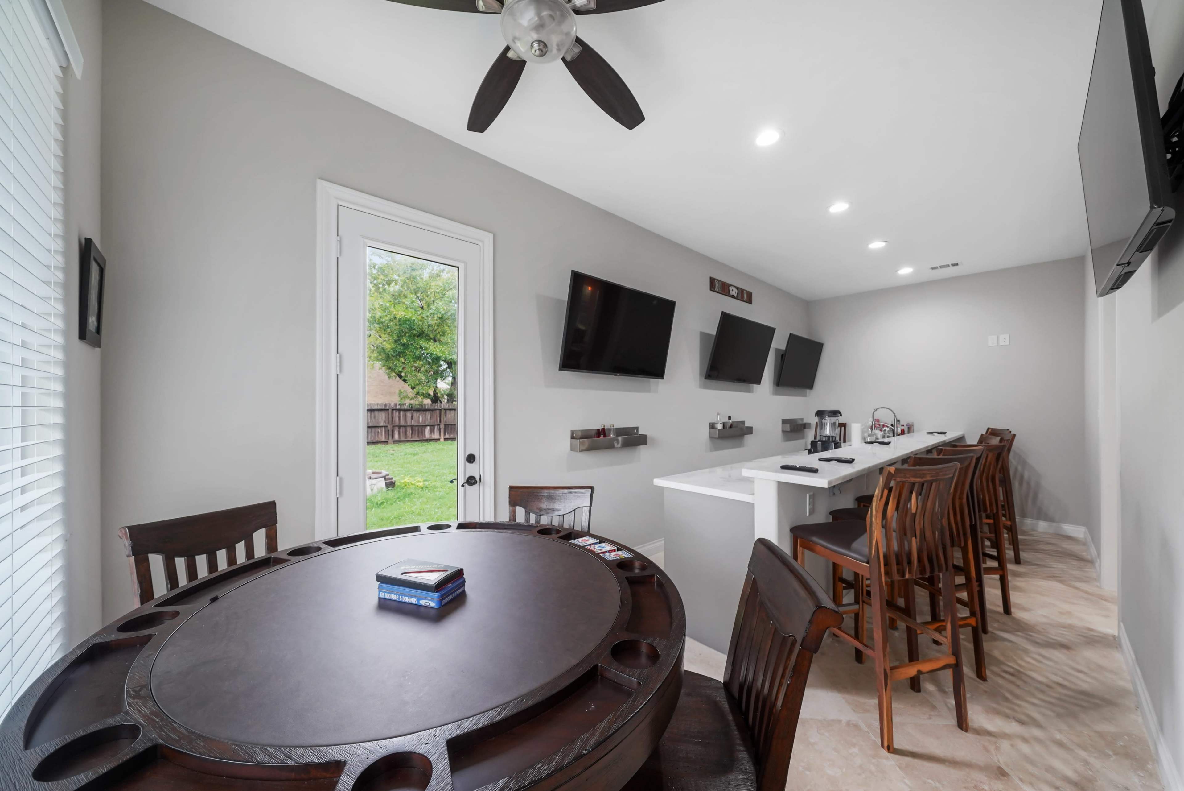 A modern indoor space featuring a round dining table, bar seating area with high chairs, and multiple wall-mounted screens.