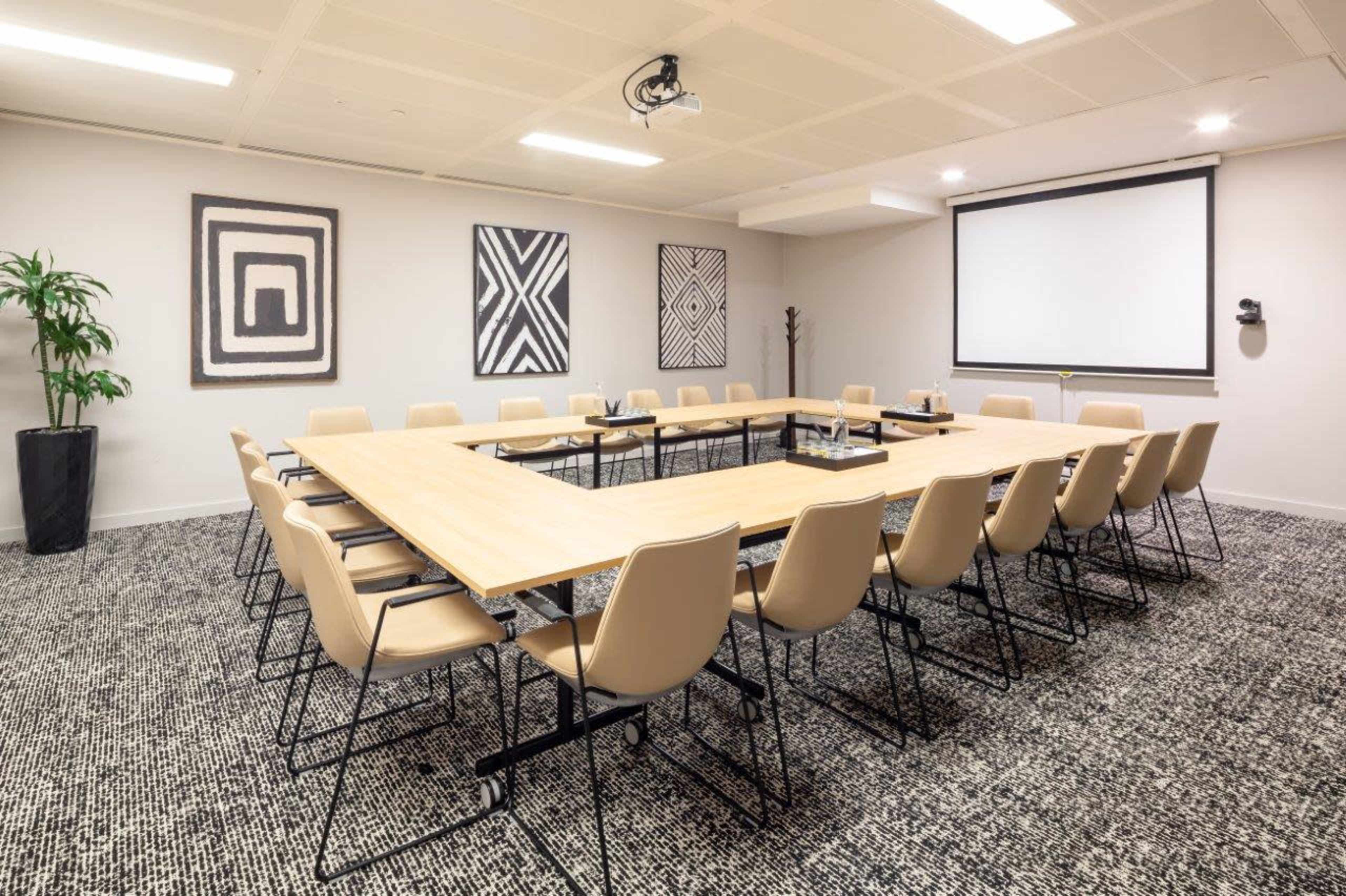 A rectangular conference room is set up with a large wooden table surrounded by beige chairs, featuring artwork on the walls and a projector screen at one end.