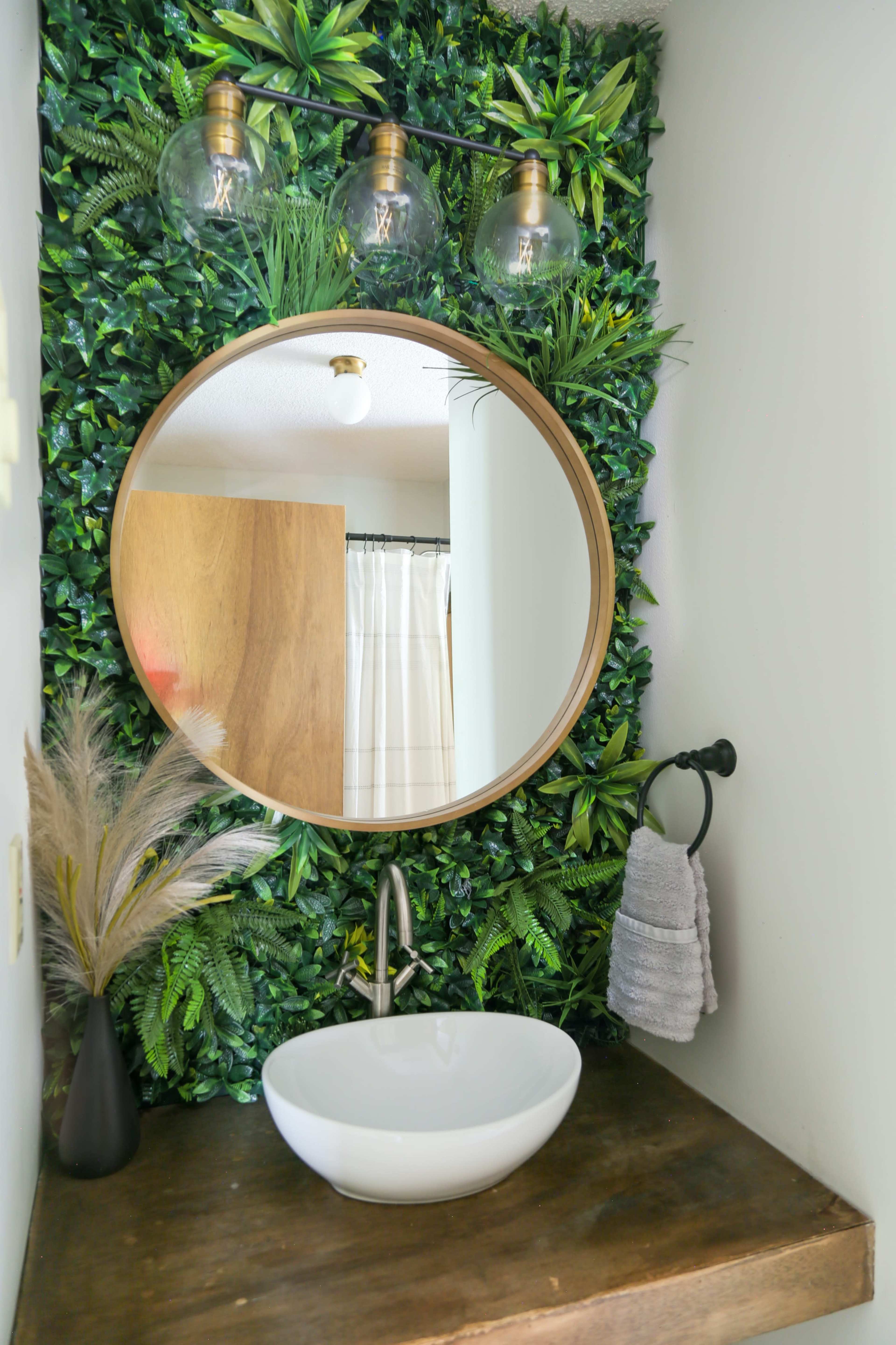 A modern bathroom features a round mirror above a white basin sink, surrounded by a leafy green wall and a wooden countertop.