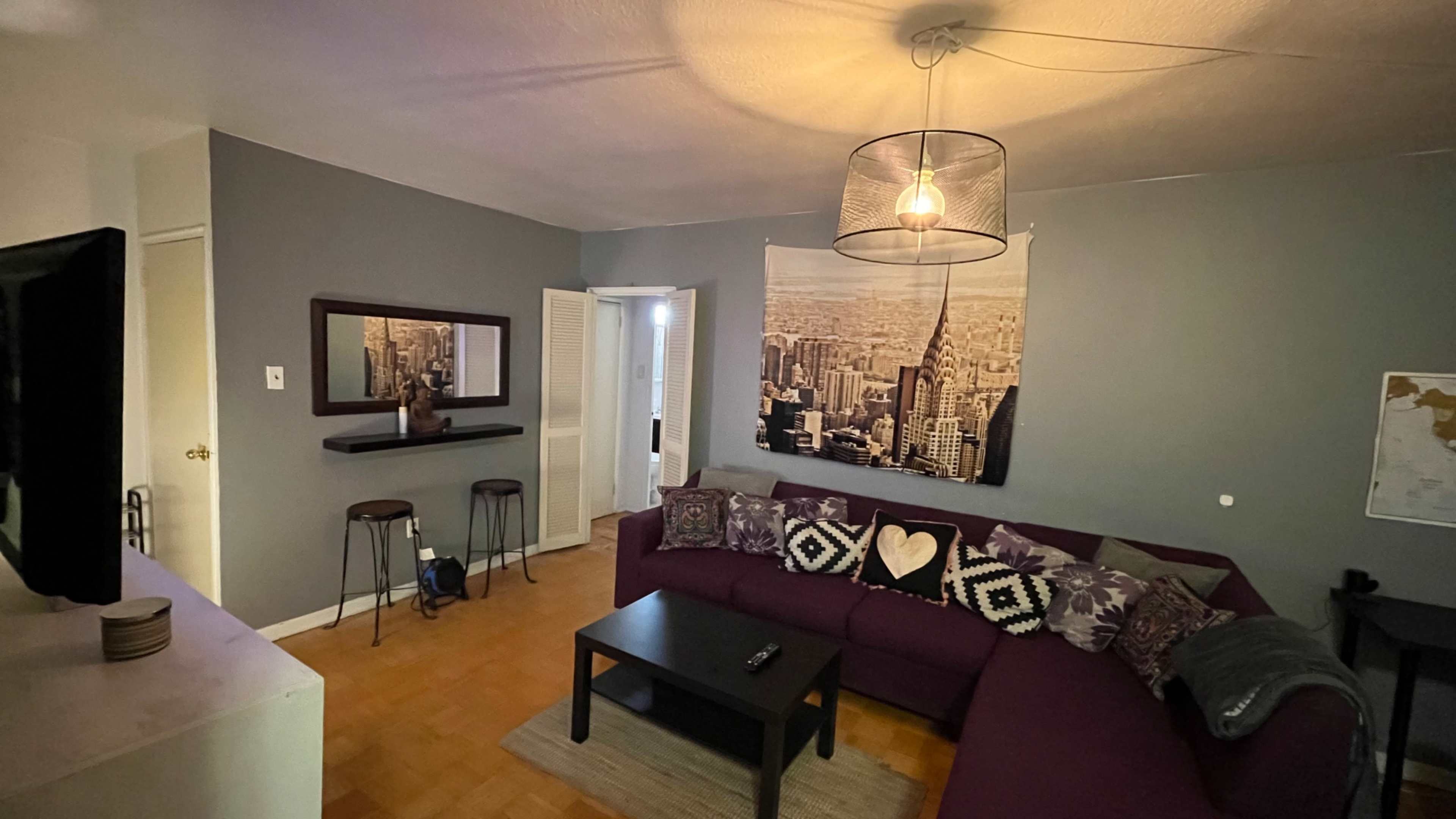 A cozy living room features a purple sectional sofa, a black coffee table, and a wall-mounted cityscape photograph.