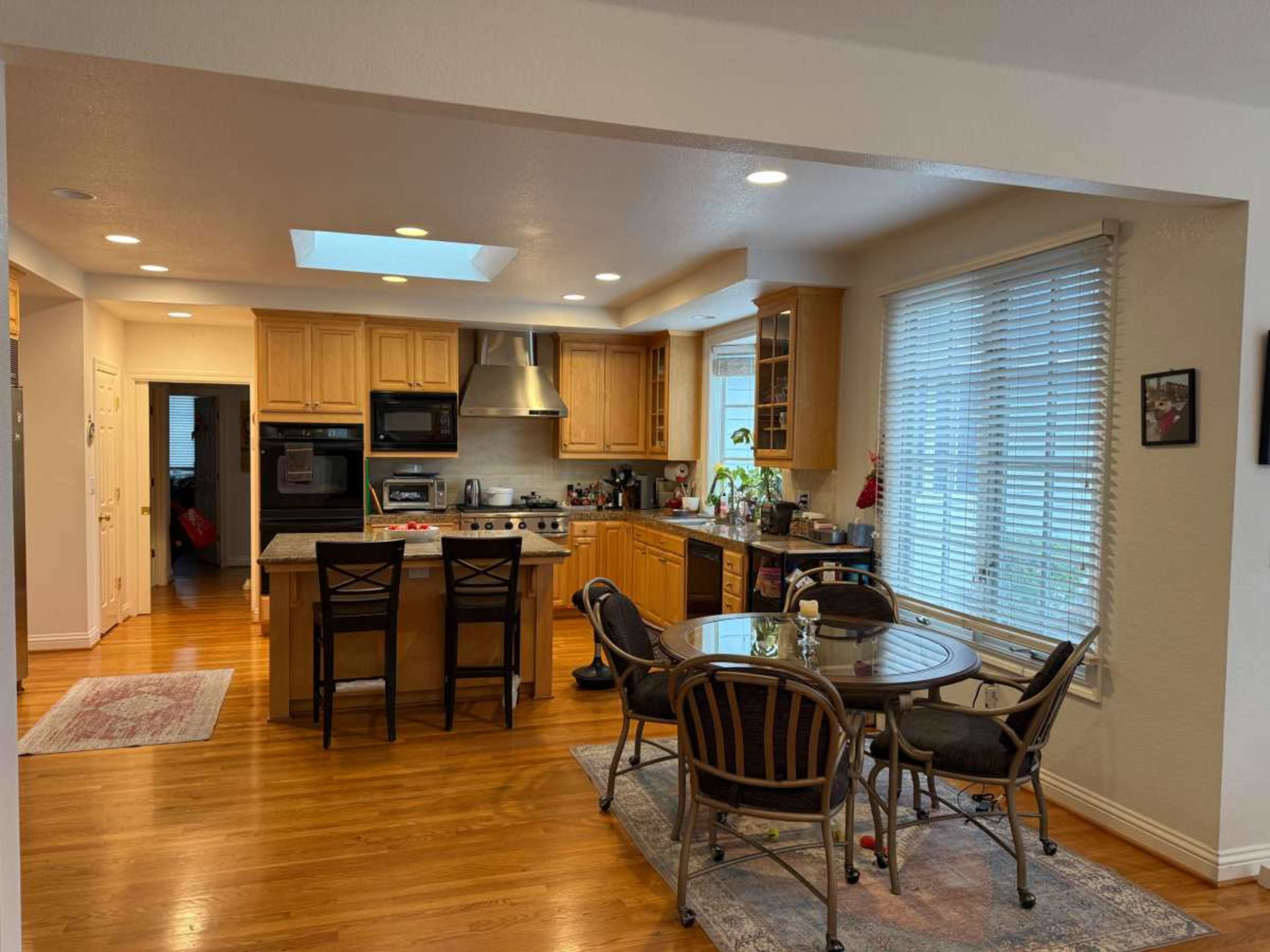 The image depicts a modern kitchen with wooden cabinetry, a central island, and a dining area featuring a round table and chairs, all illuminated by natural light from large windows.