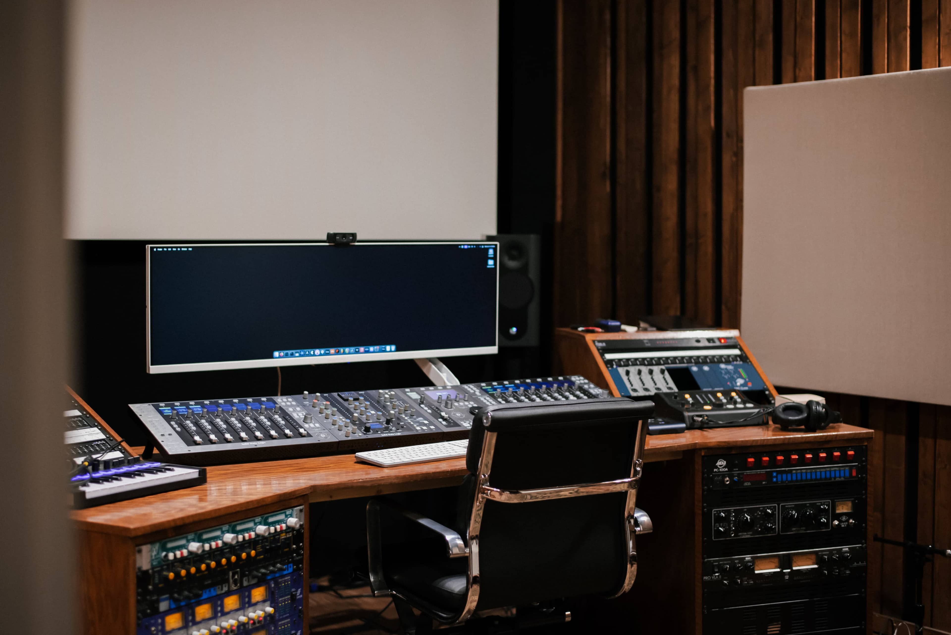 The image shows a modern audio mixing studio with a large computer monitor, a digital mixing console, and various audio equipment arranged around a desk.
