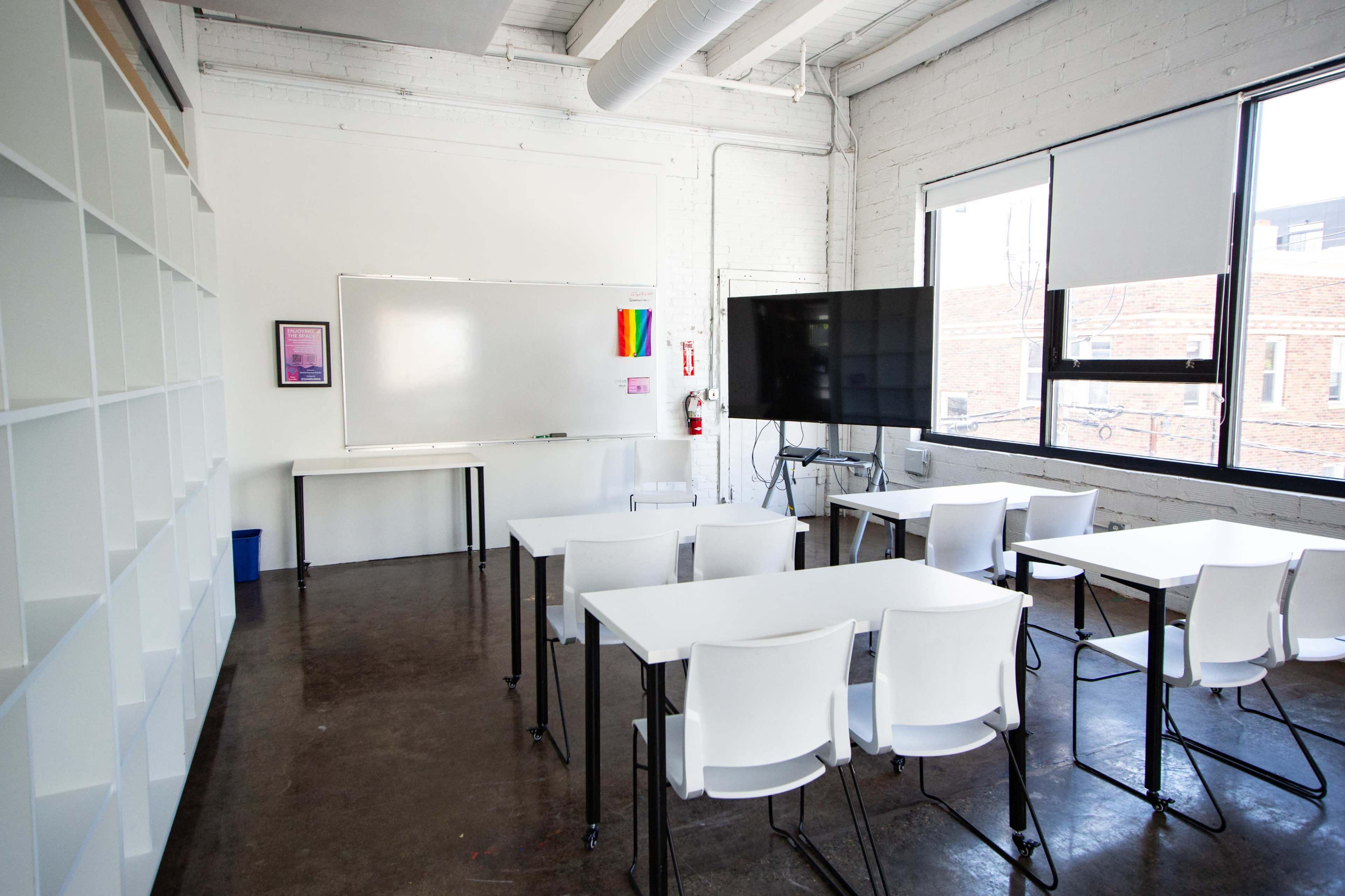 The image shows a modern classroom with white tables and chairs, a large screen on a stand, a whiteboard, and shelves in the background.