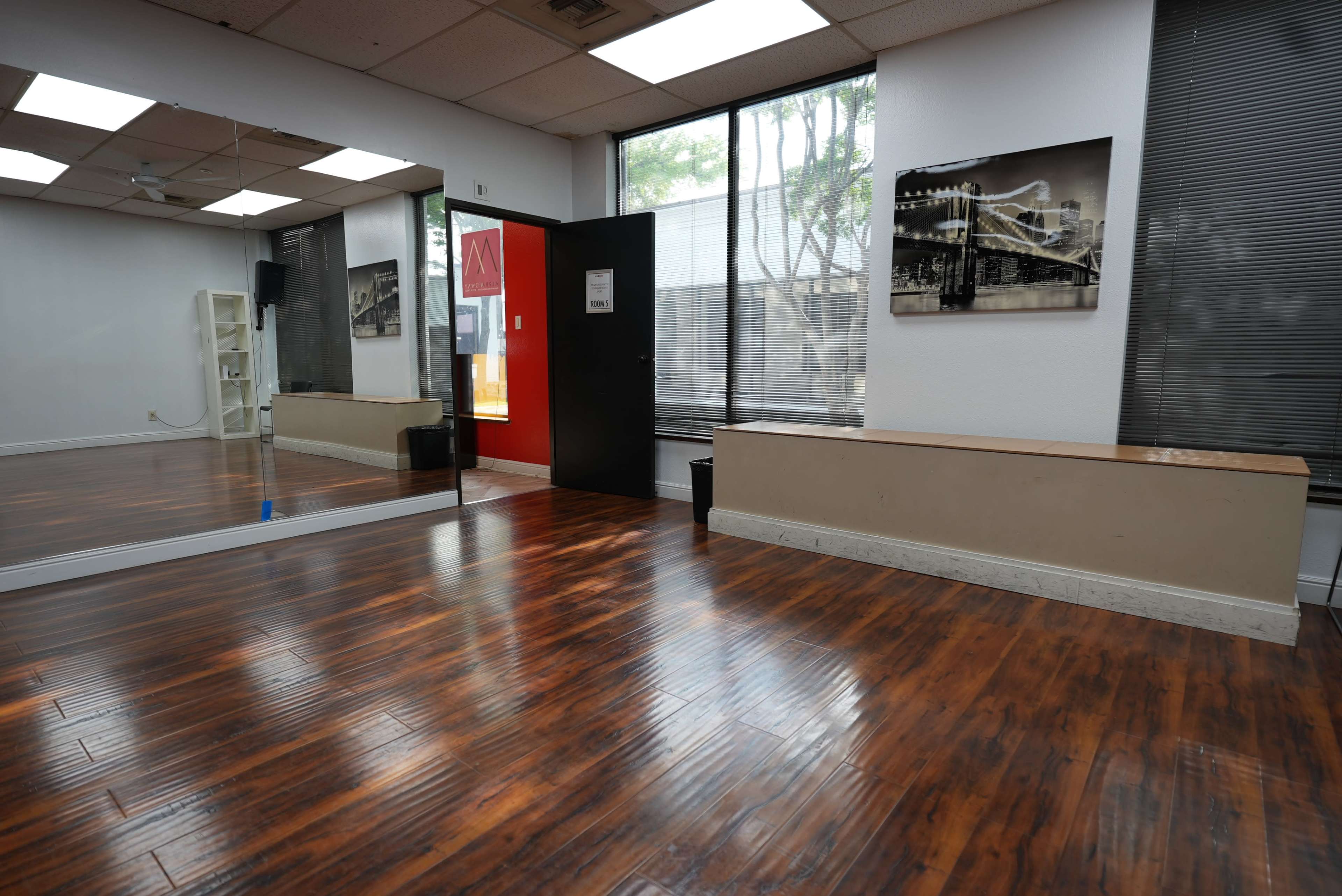 The image shows an empty studio with wooden flooring, large windows, a mirrored wall, and a bench along one side.
