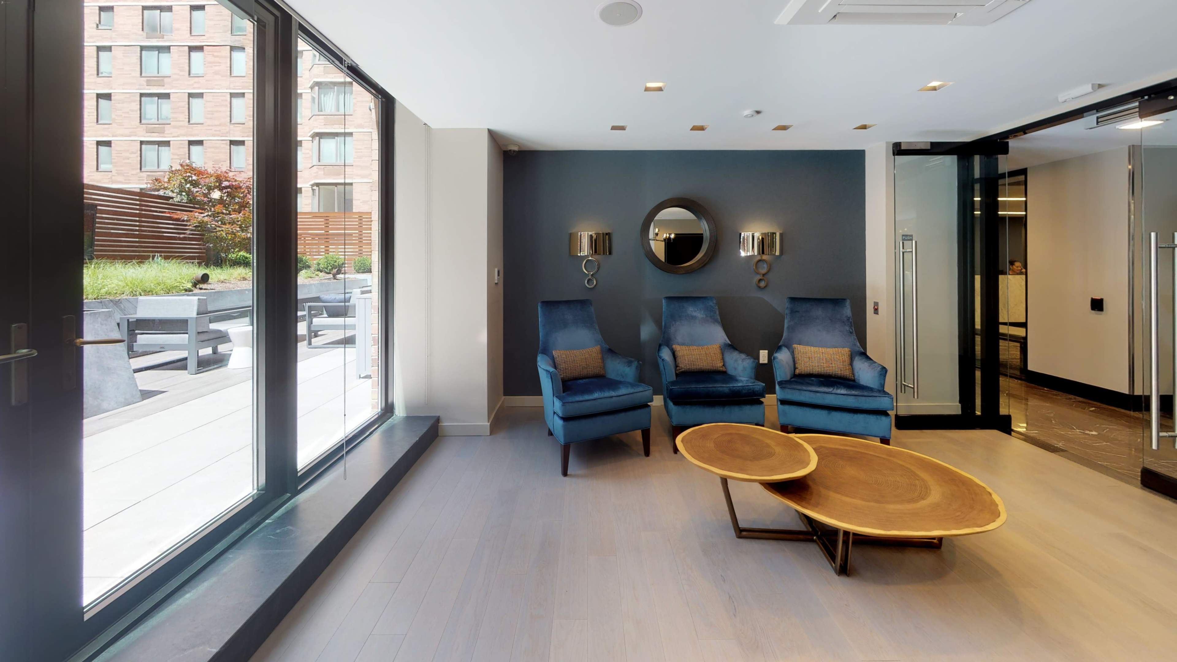 A modern lounge area features two blue chairs, a round wooden coffee table, and large windows that overlook an outdoor patio.