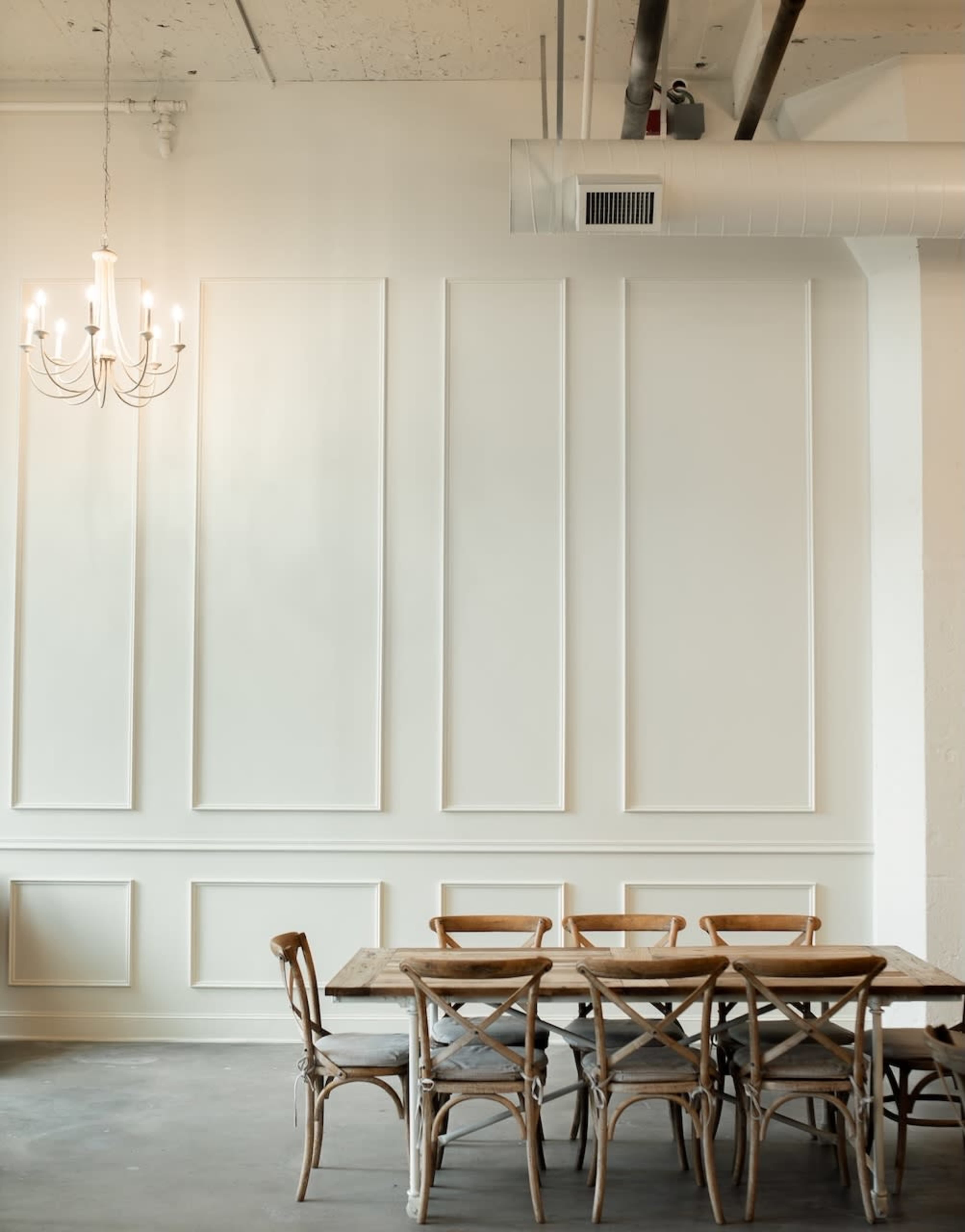 A wooden dining table with matching chairs is positioned beneath a chandelier in a room featuring white paneled walls.