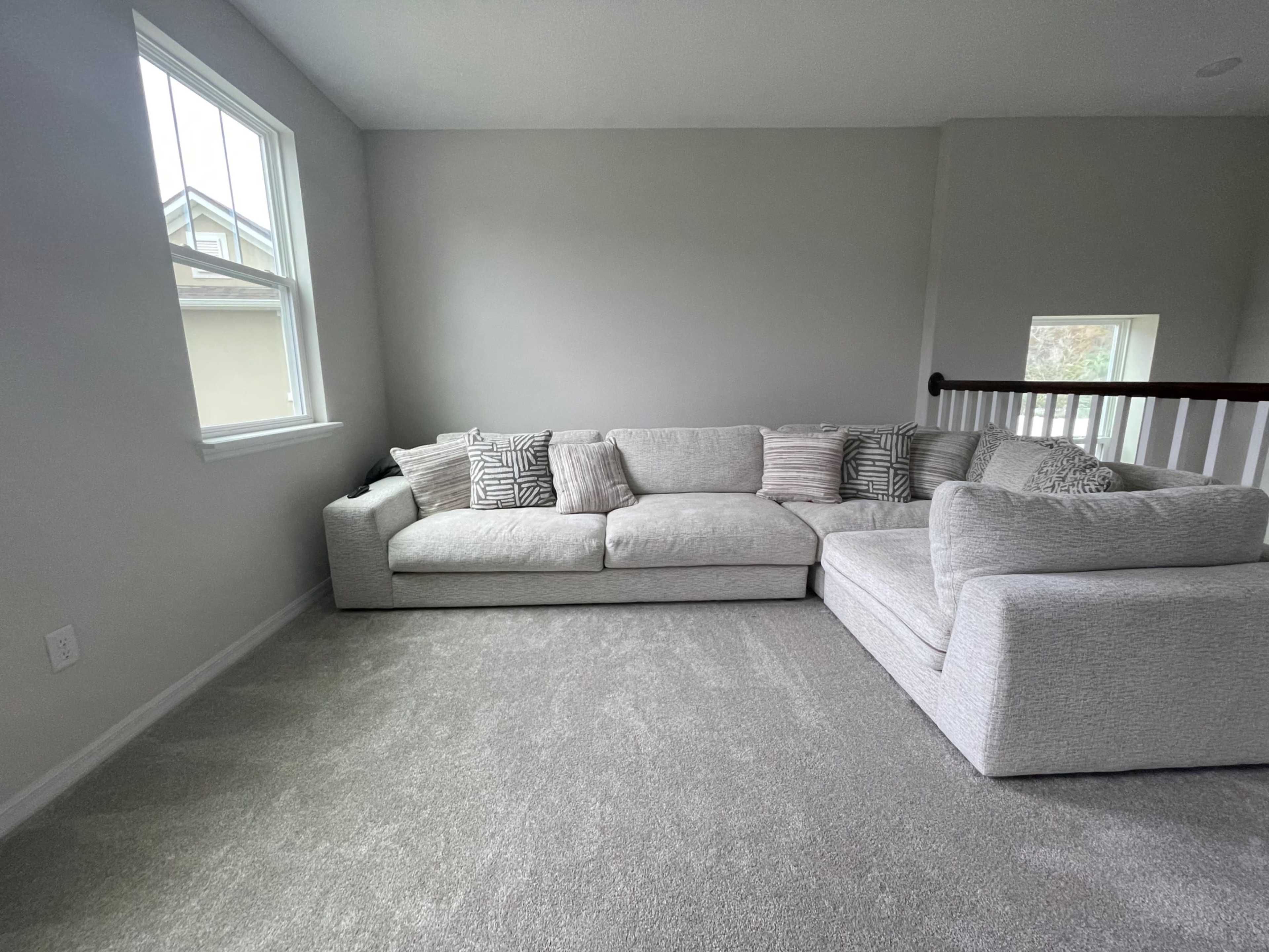 A spacious living area features a light-colored sectional sofa against a grey carpet with large windows allowing natural light.