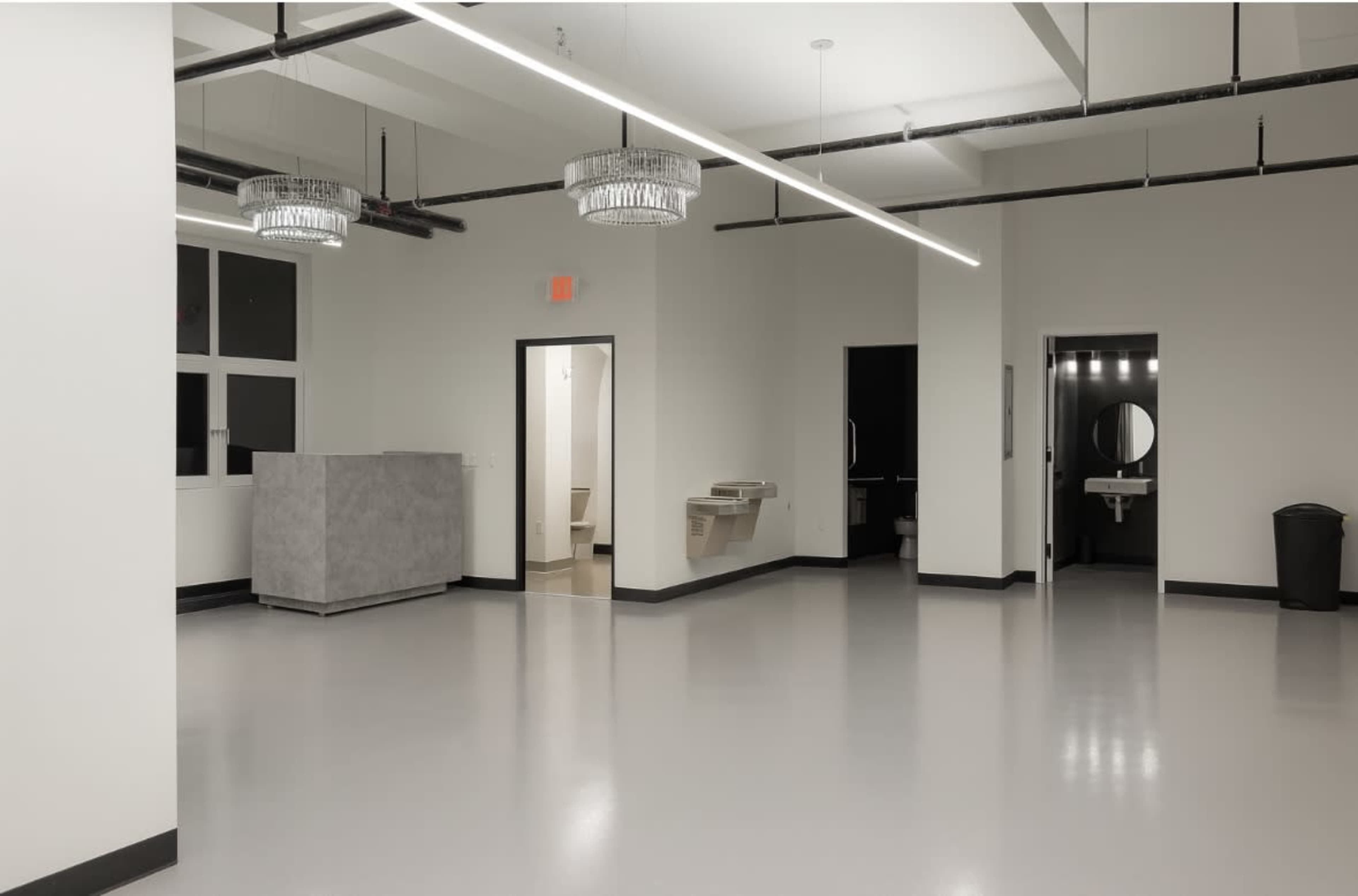 The image shows a large, empty, modern interior space with polished gray floors, minimalist design, and two doorways leading to restrooms.