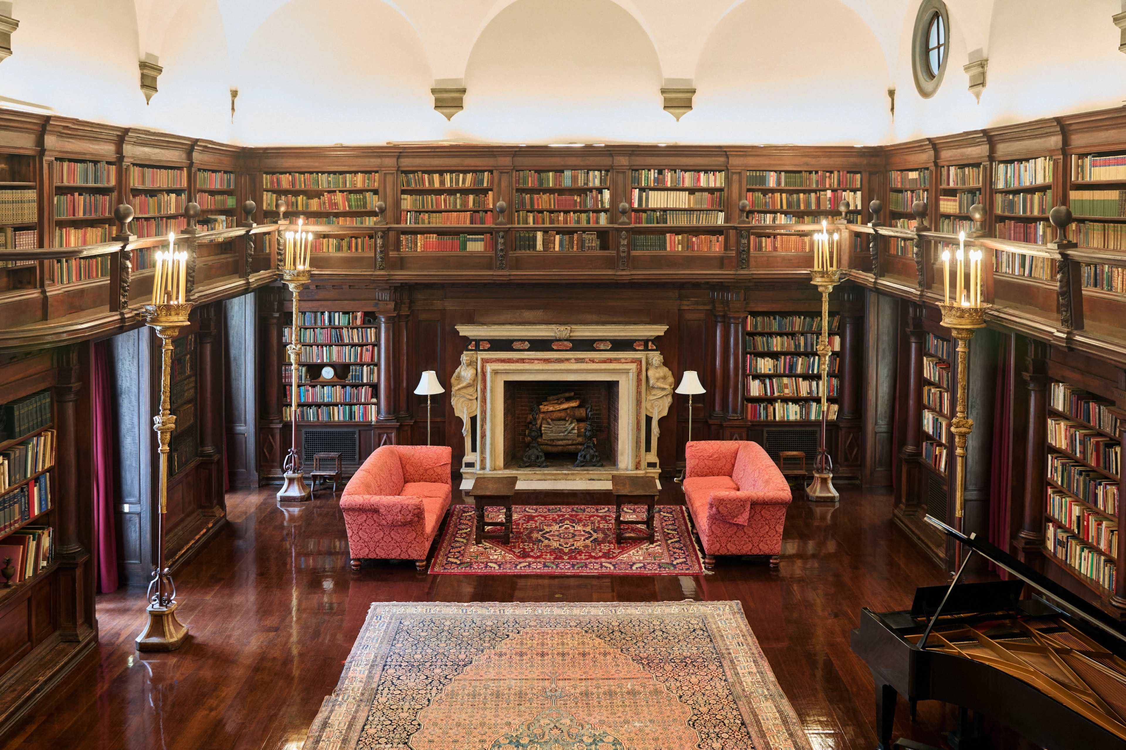 The image shows a spacious, elegantly furnished library with large bookshelves, a fireplace, two armchairs, a coffee table, and a grand piano.