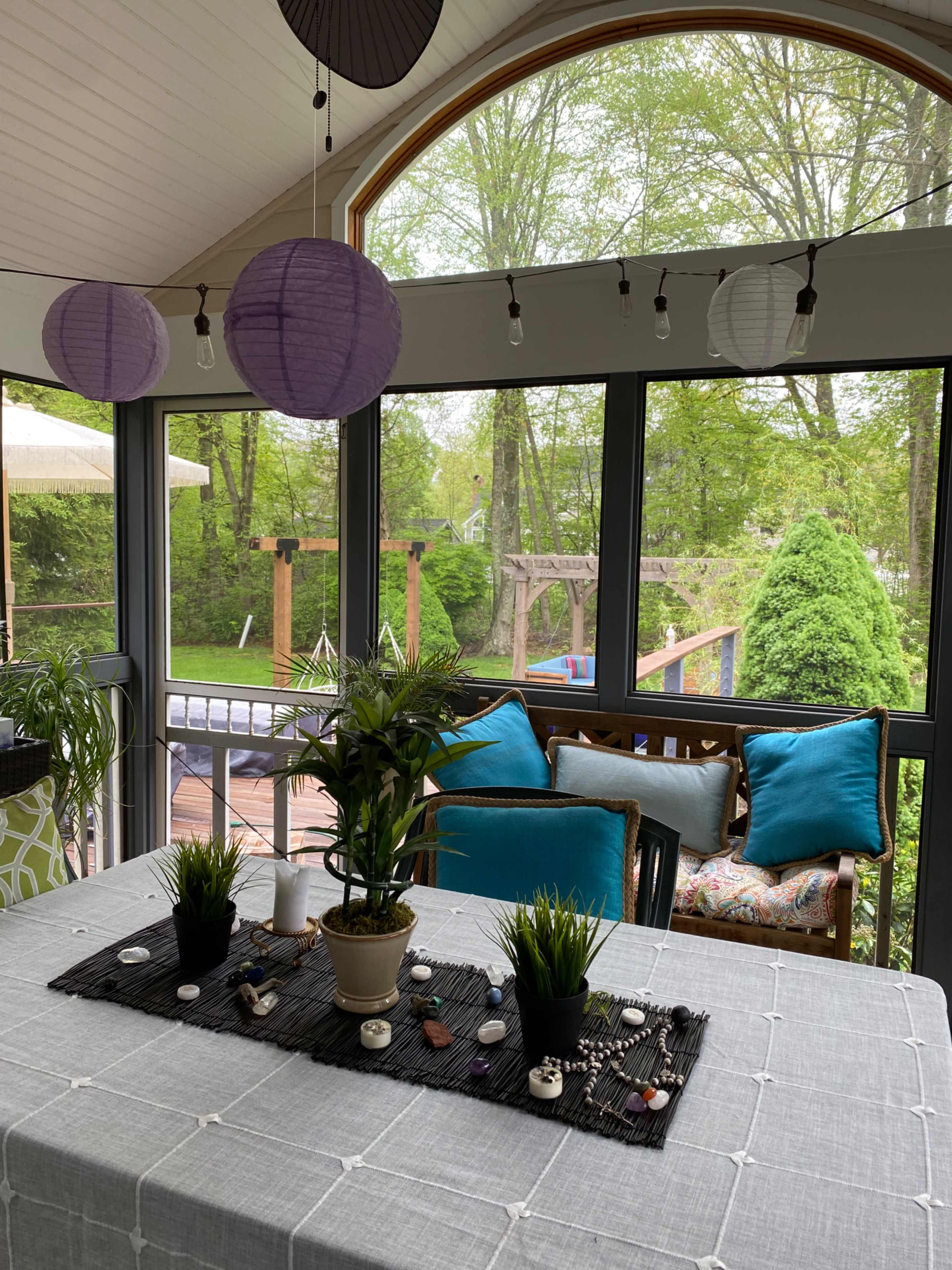 A bright sunroom features a dining table adorned with plants and decorative stones, surrounded by large windows overlooking a green outdoor space.