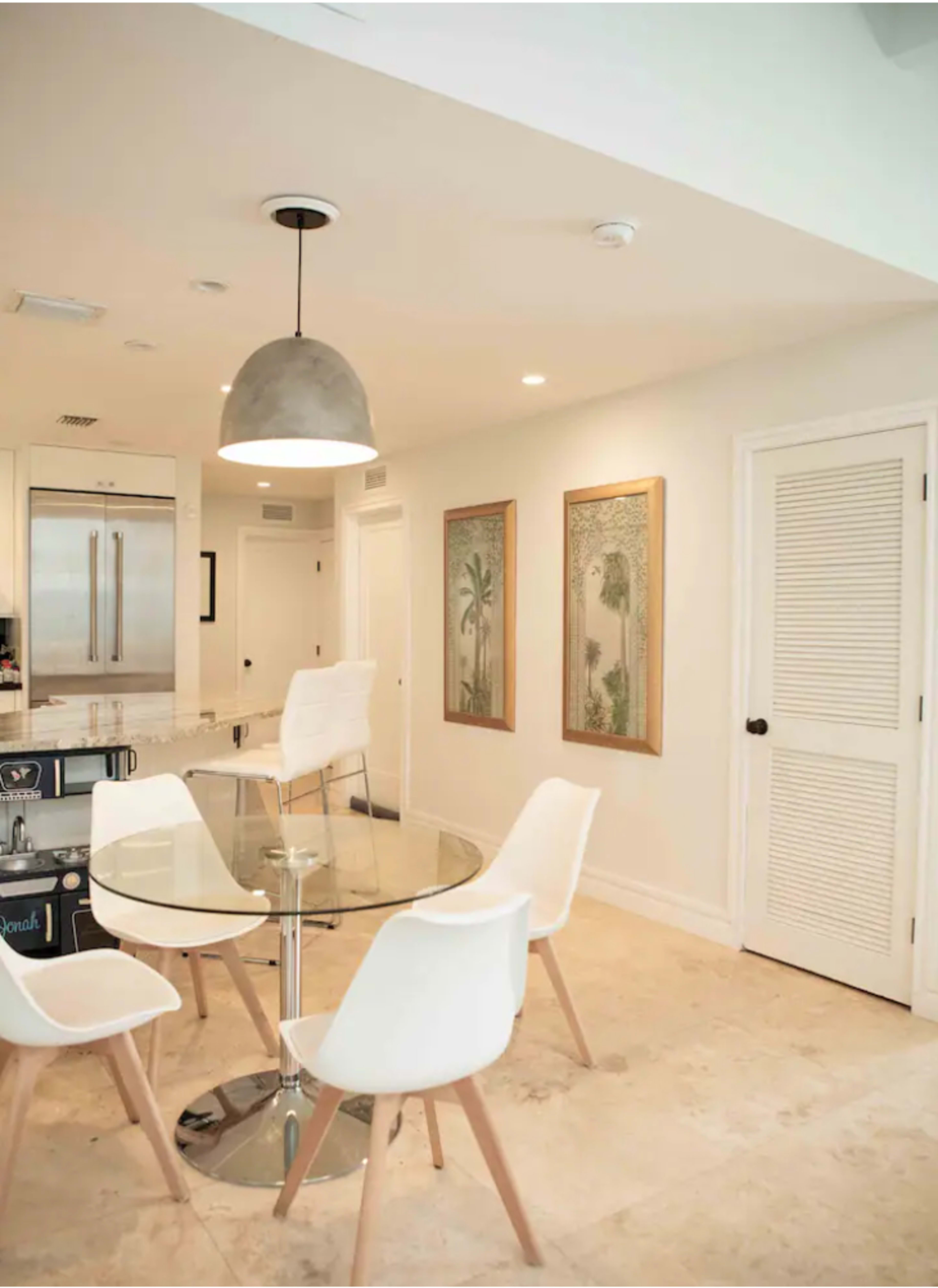 A modern kitchen features a round glass dining table surrounded by four white chairs, with framed artwork on the walls and a pendant light above.