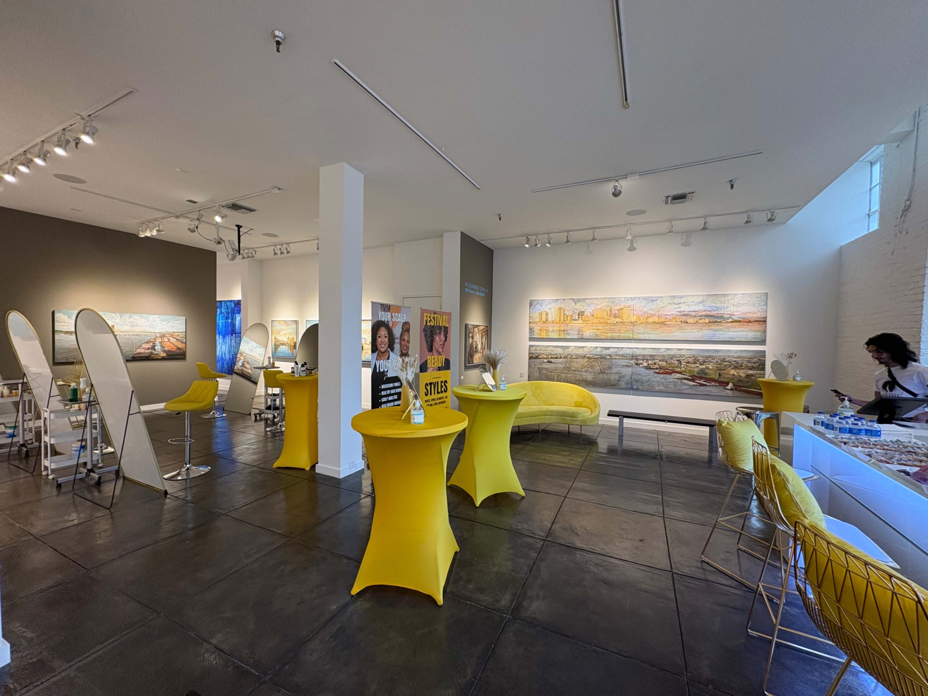 The image shows a modern art gallery featuring bright yellow furniture, abstract paintings on the walls, and a variety of seating arrangements.
