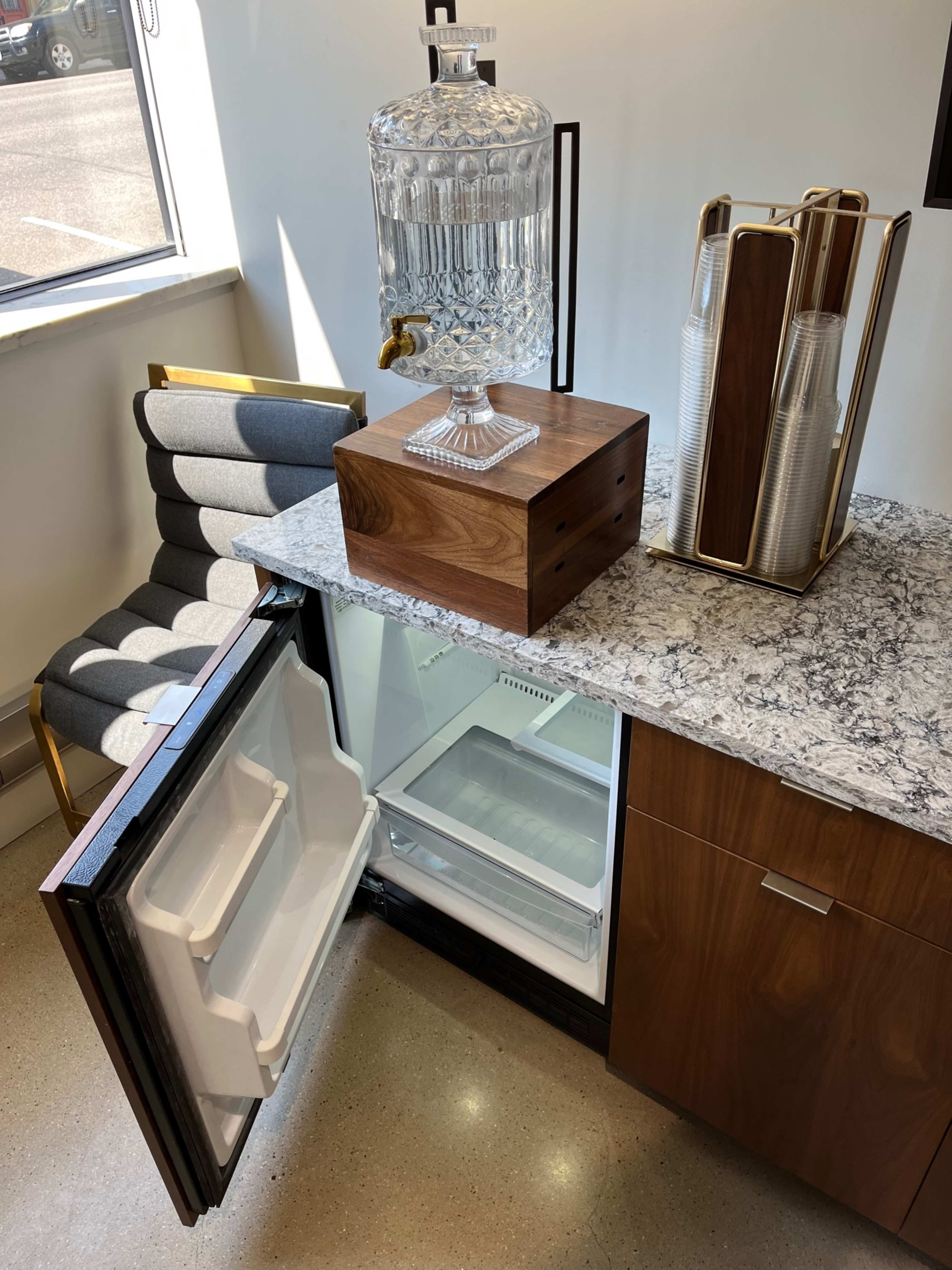 An open refrigerator with a decorative water dispenser and cups on top of a countertop, alongside a modern chair.