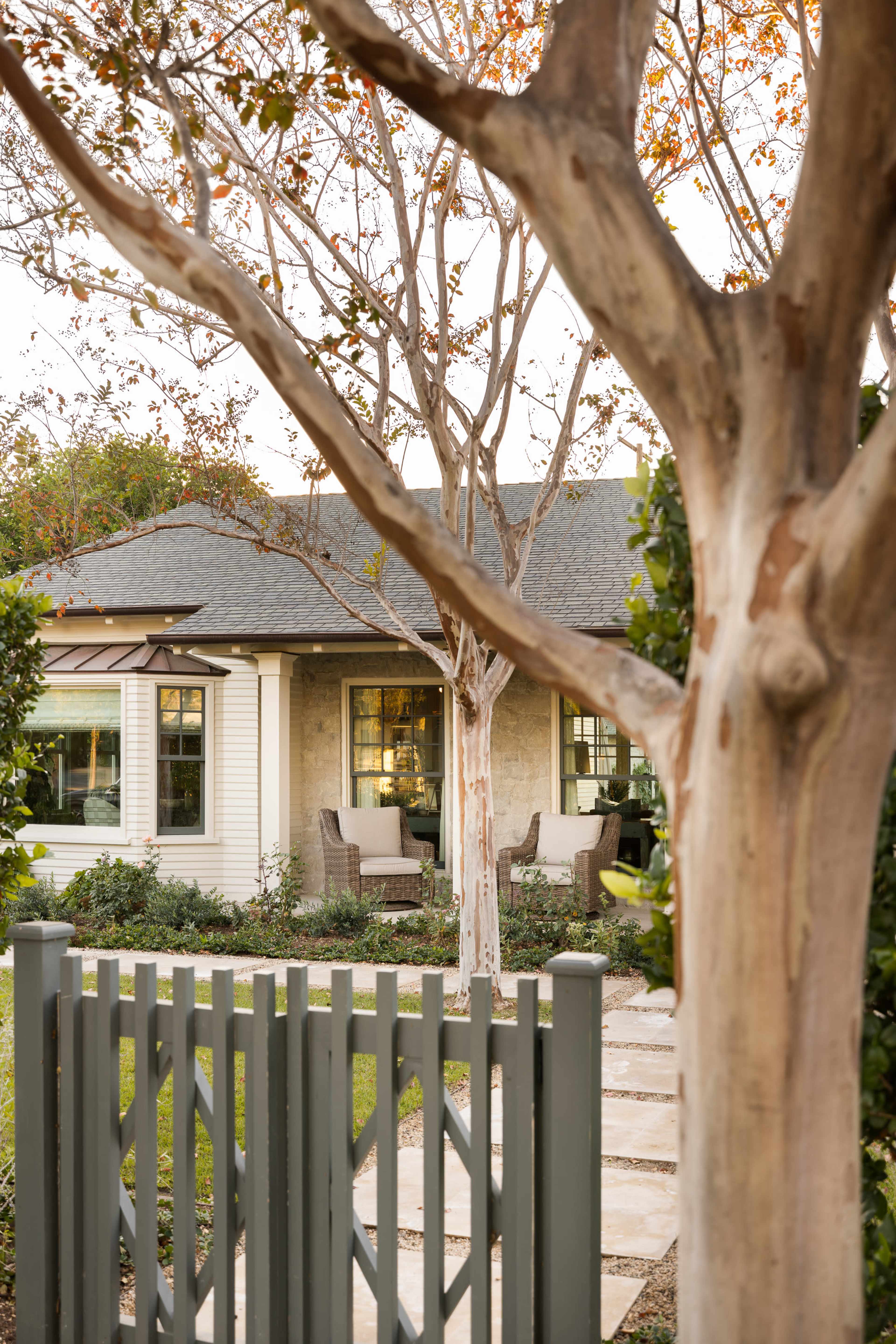 A wooden gate opens to a pathway lined with stones leading to a house with large windows and outdoor seating, framed by trees.