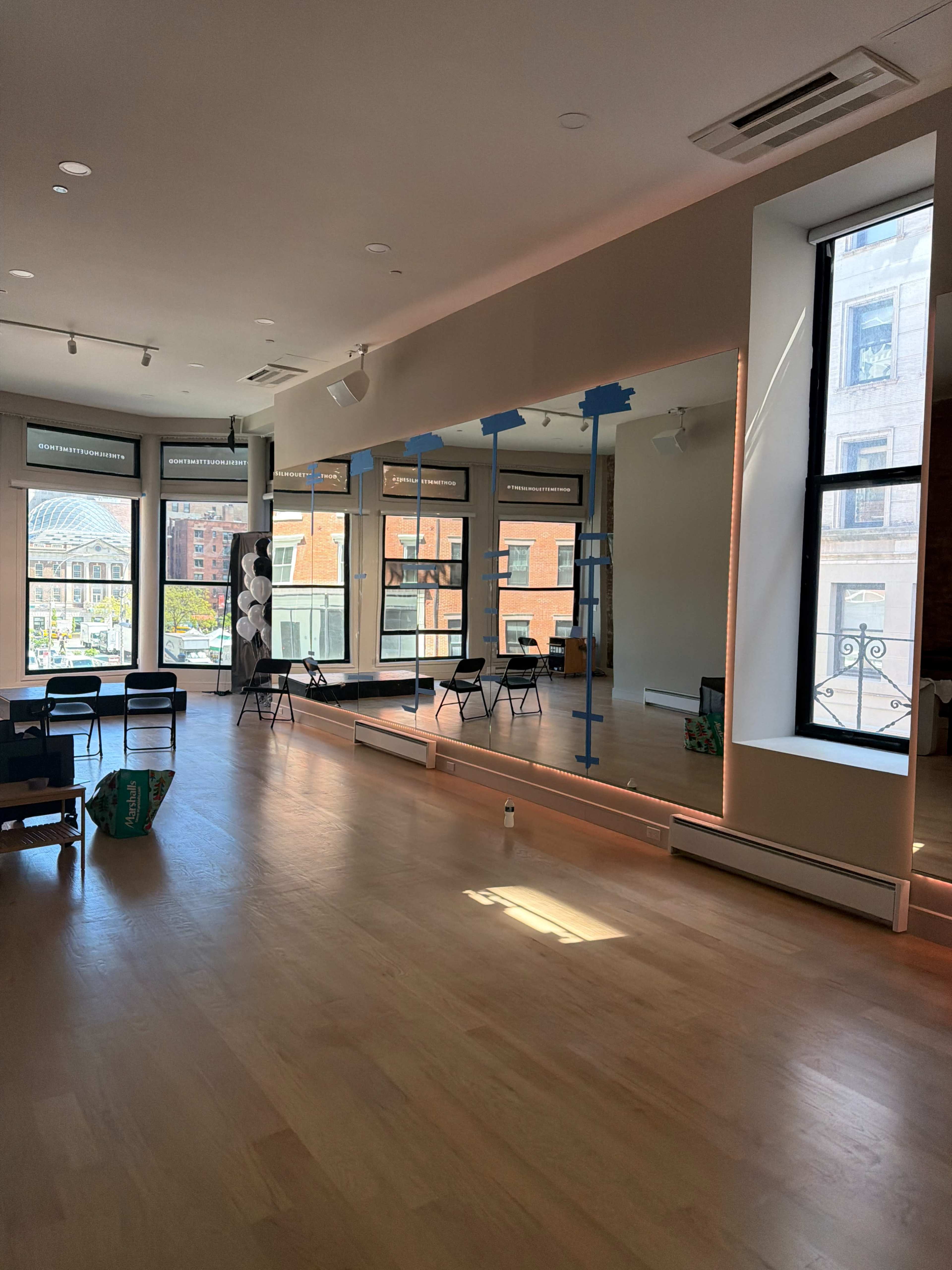 The image shows an empty dance studio with large mirrors, wooden floors, and natural light streaming through windows.