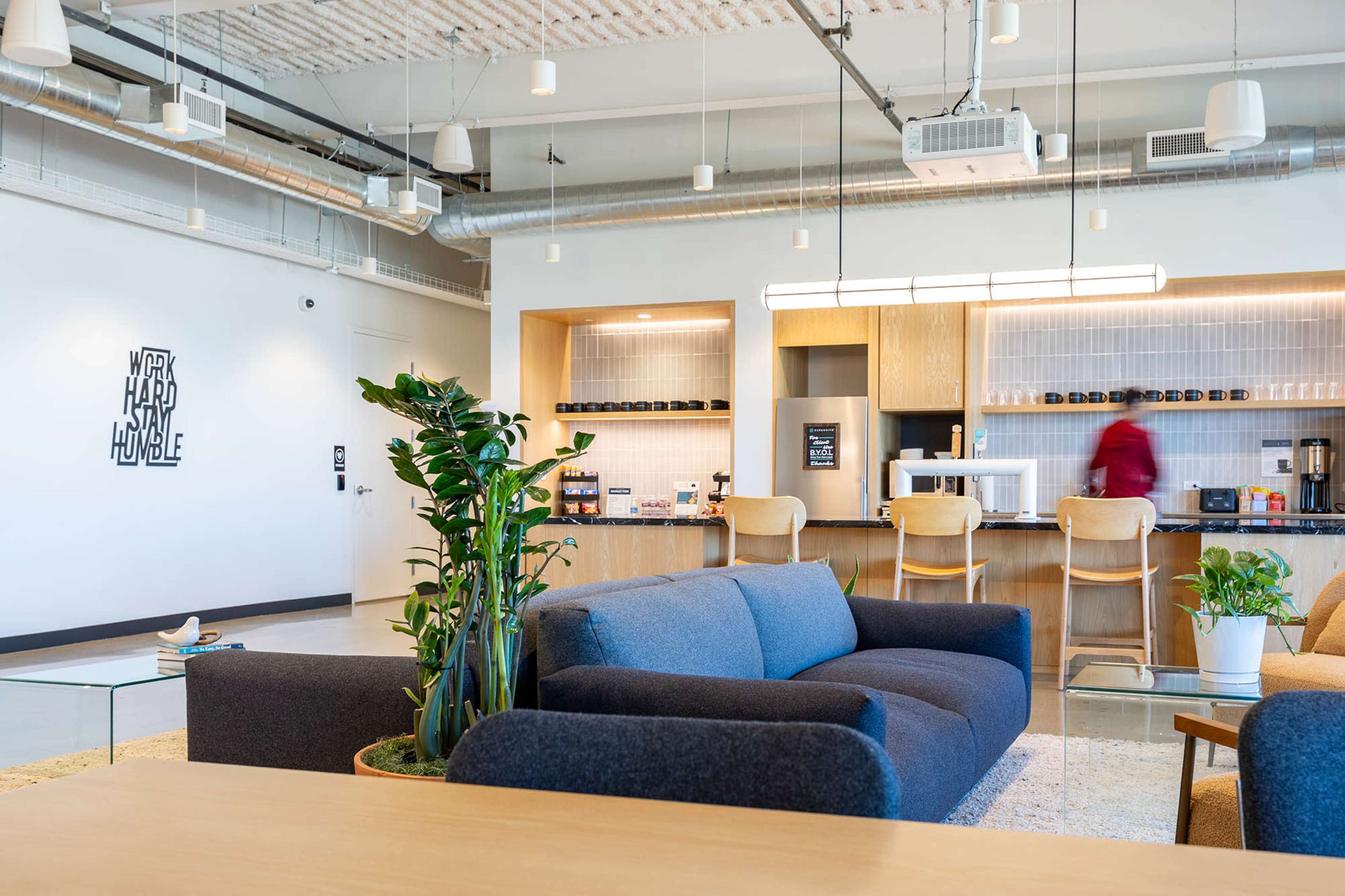 The image shows a modern office lounge with a blue sofa, wooden furniture, plants, and a beverage station in the background.
