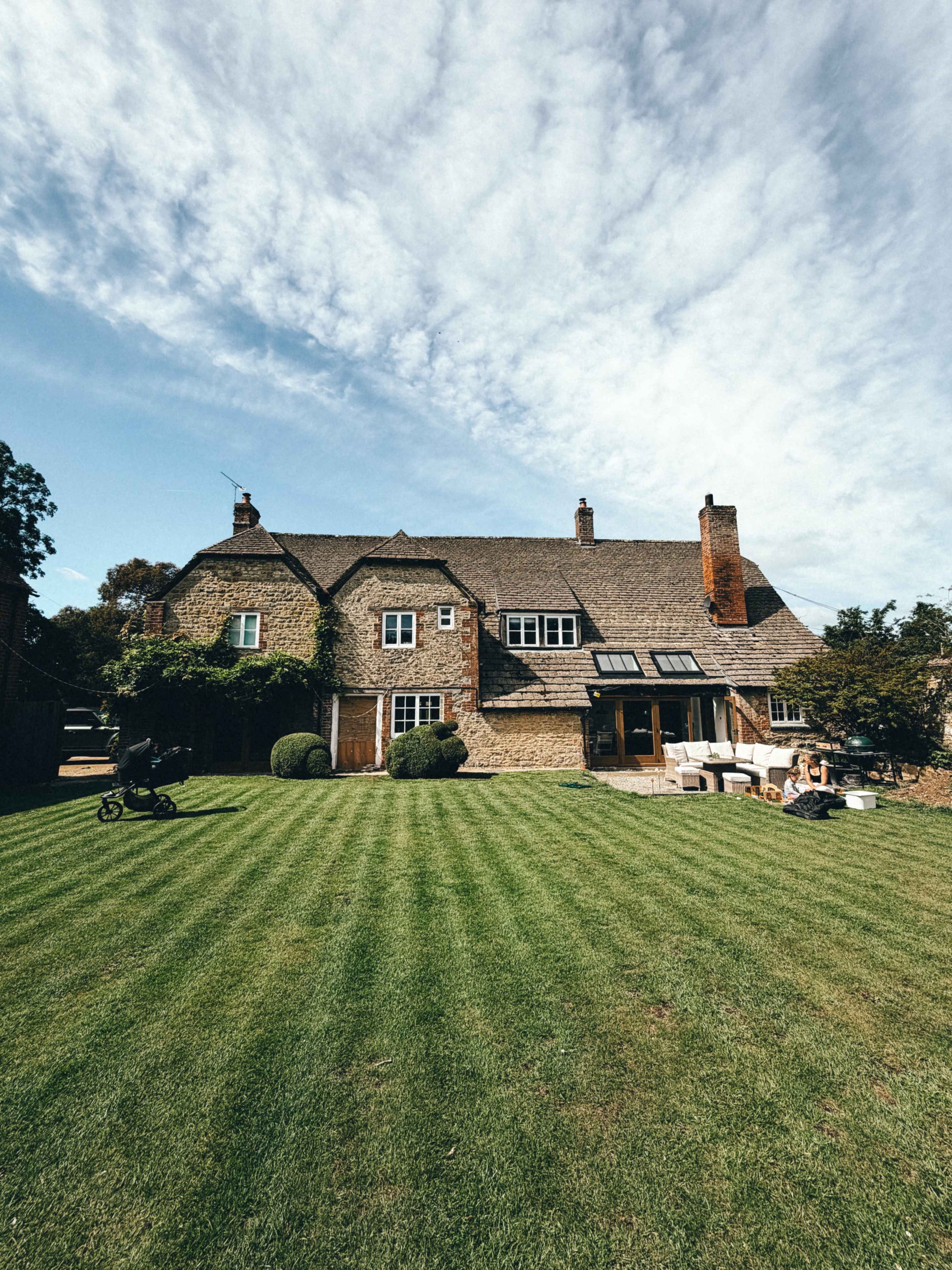 A stone house with a sloped roof and large windows sits on a neatly manicured lawn under a partly cloudy sky.