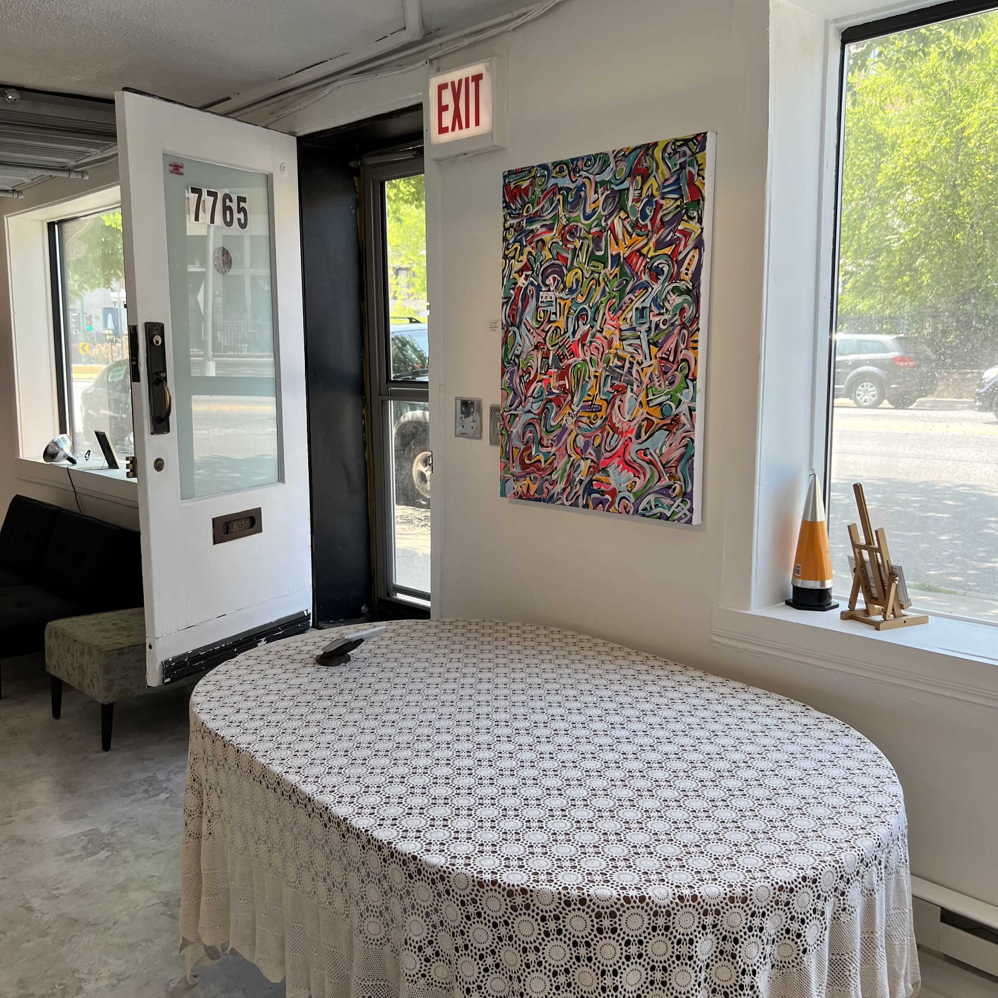 The image shows a room with a large oval table covered by a lace tablecloth, a colorful abstract painting on the wall, and an exit door leading outside.
