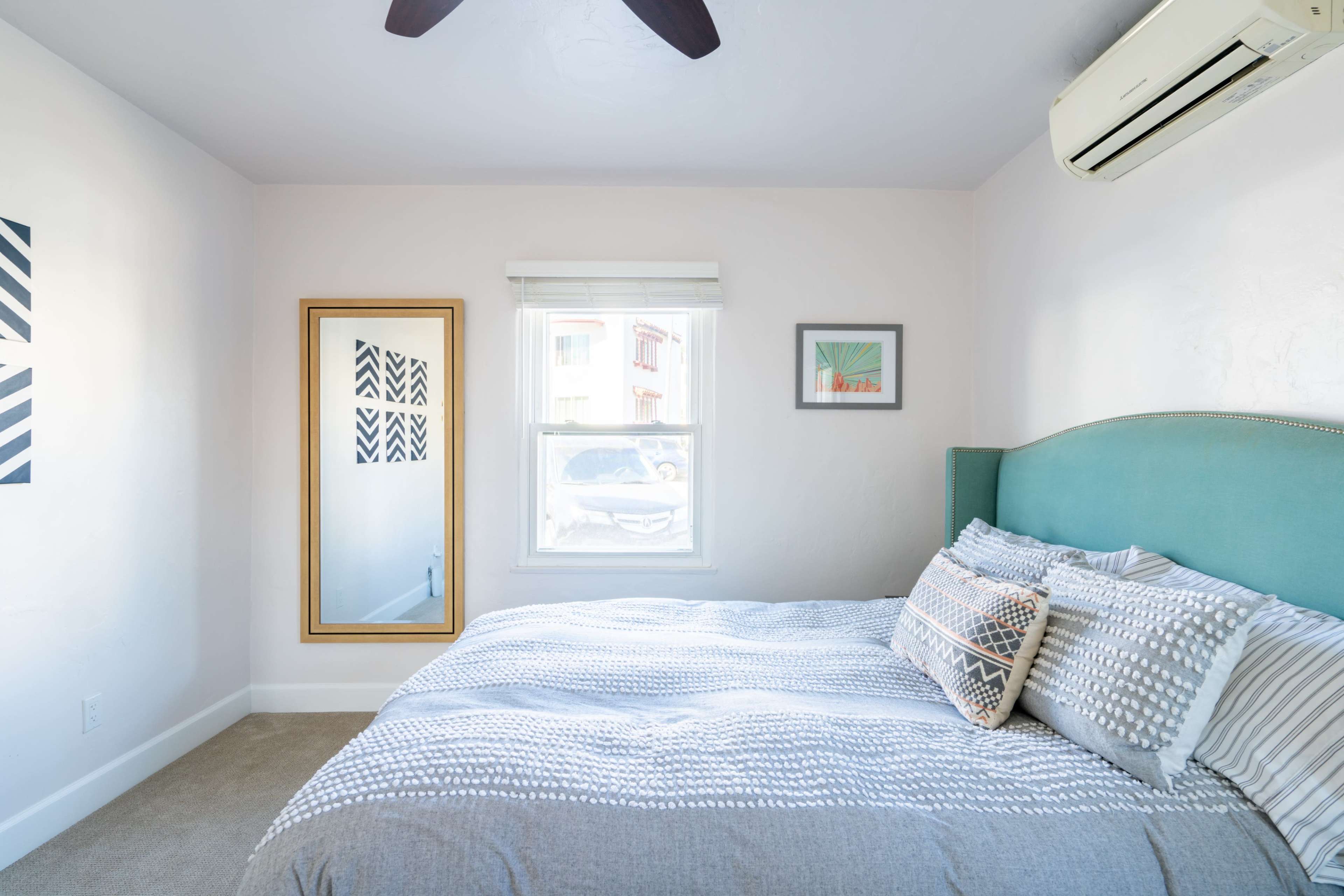 A bright bedroom features a bed with gray linens, a teal headboard, and a large mirror reflecting the room's decor.