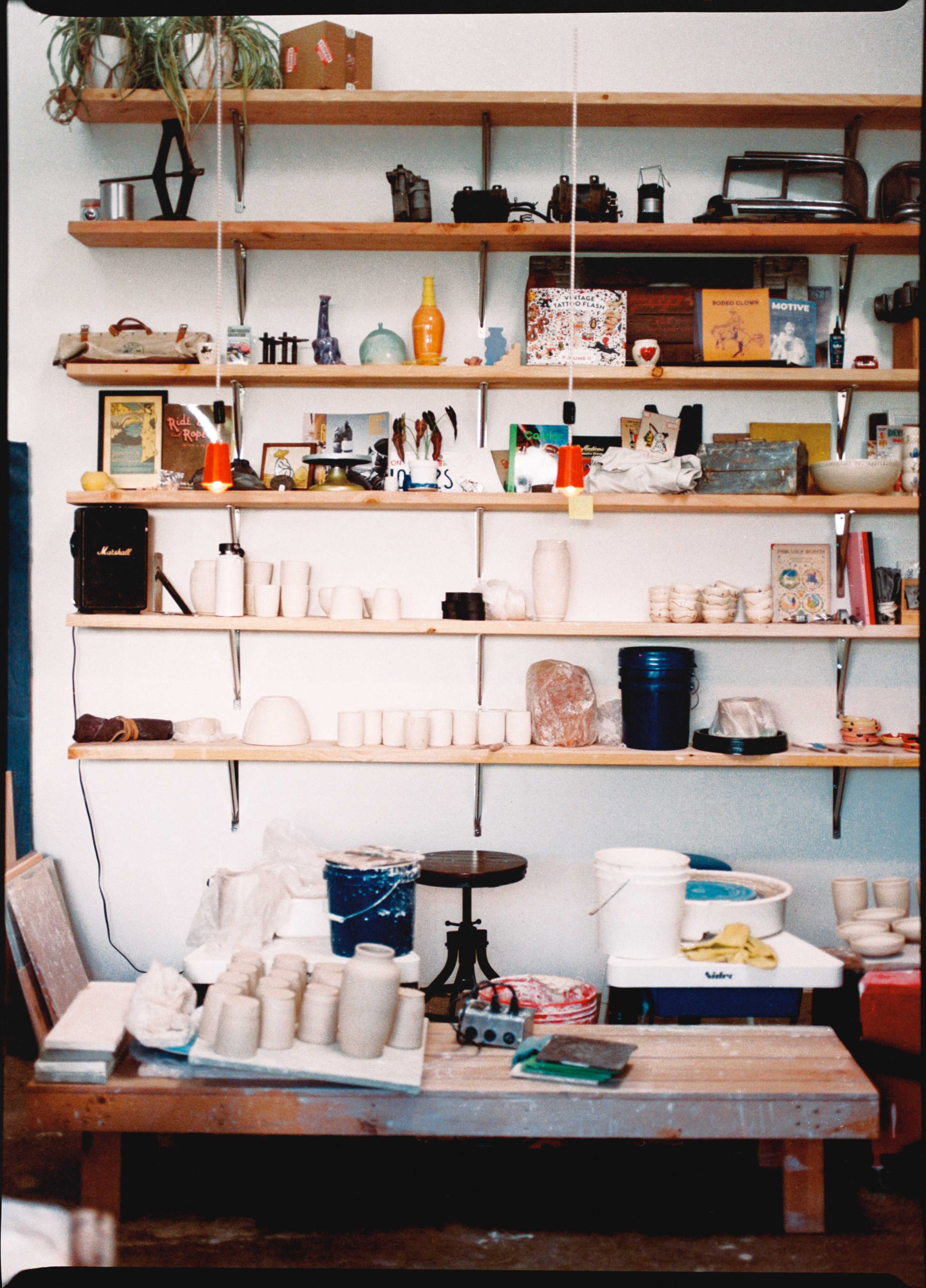 A cluttered pottery studio features shelves with various ceramics, tools, and decorative items, along with a workbench in front.