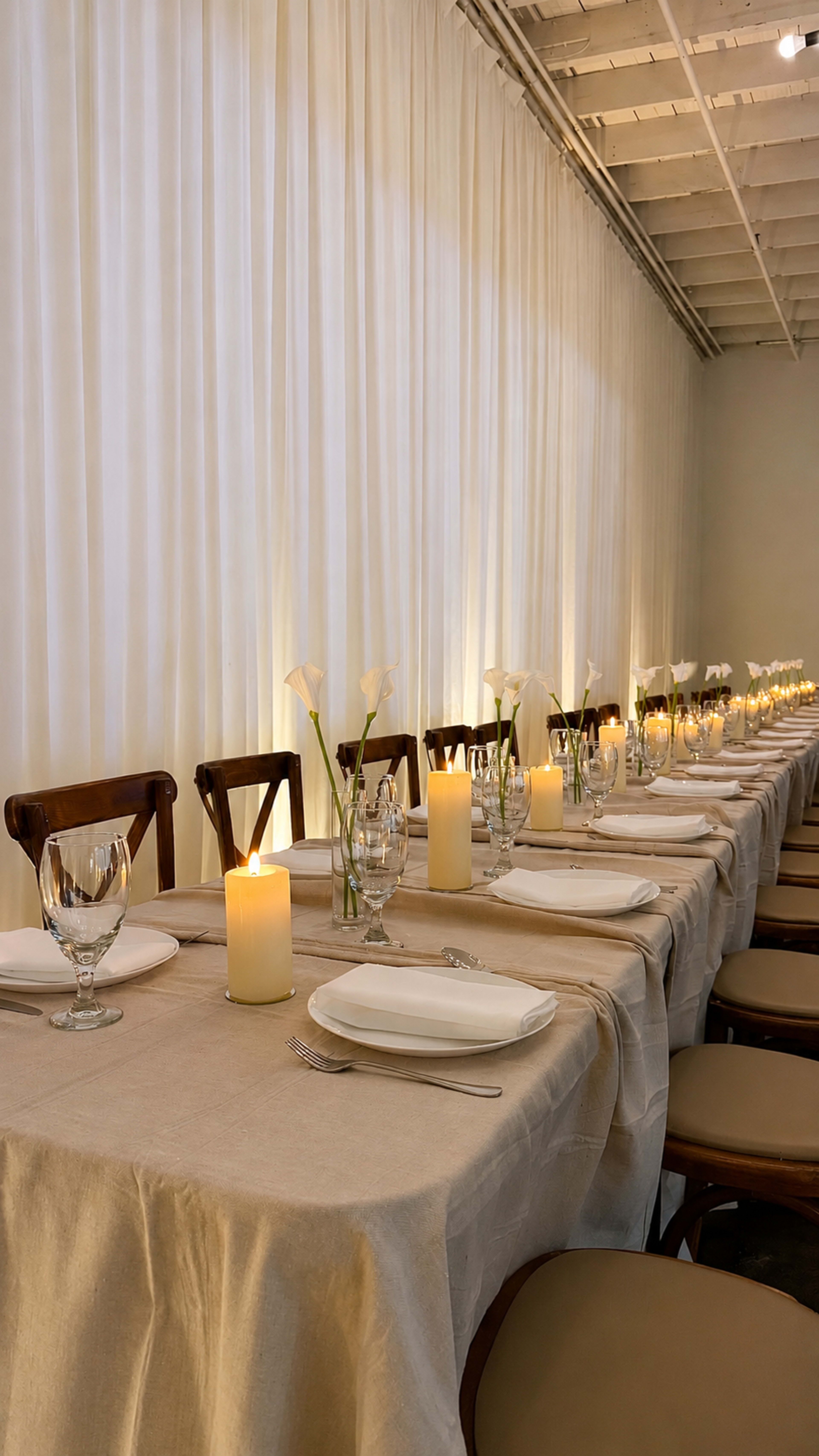 A long dining table is set with white tablecloths, glassware, and candles, surrounded by soft fabric curtains.