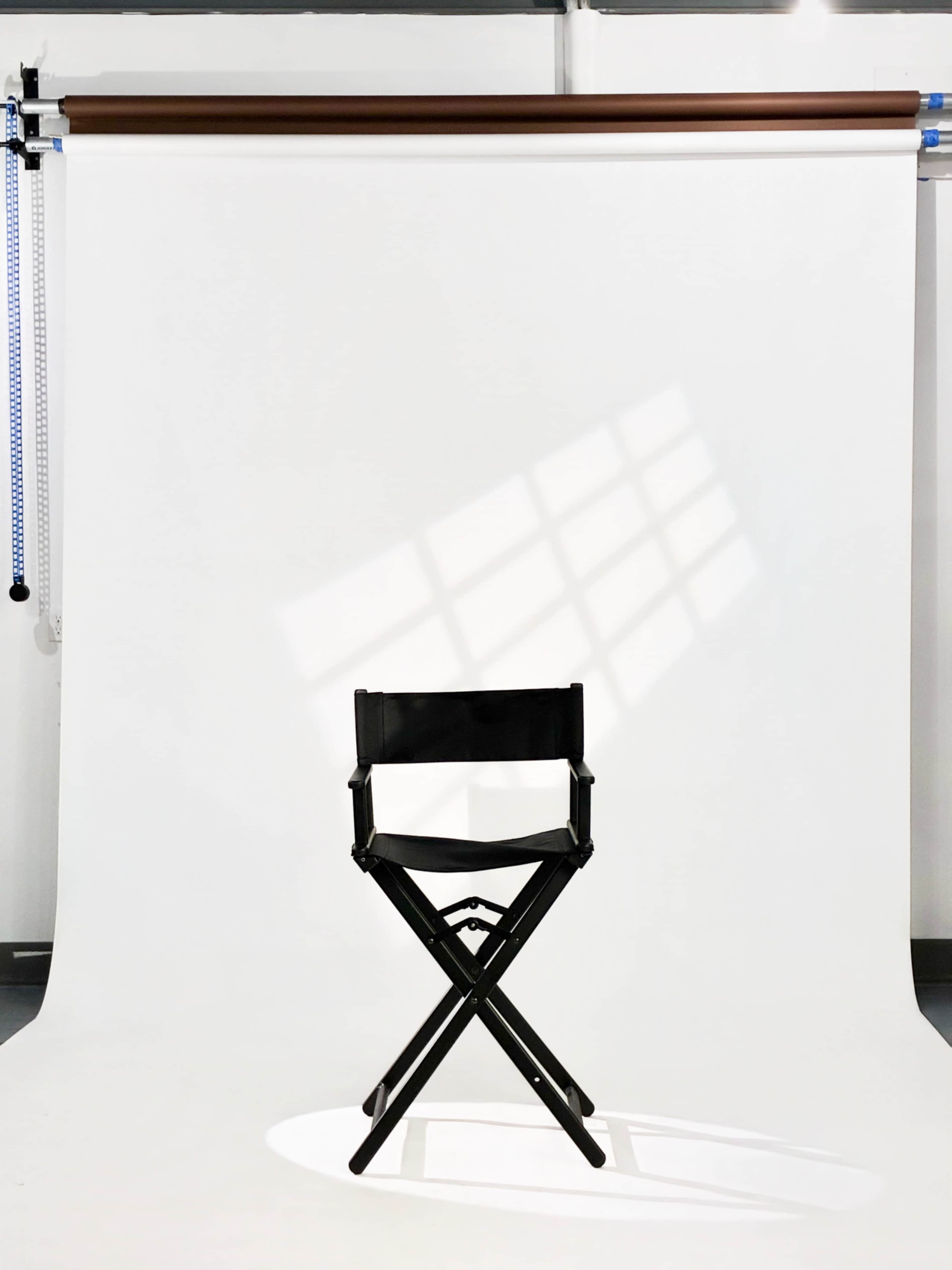 A black director's chair is positioned in front of a white backdrop with a shaft of light casting a shadow on the floor.