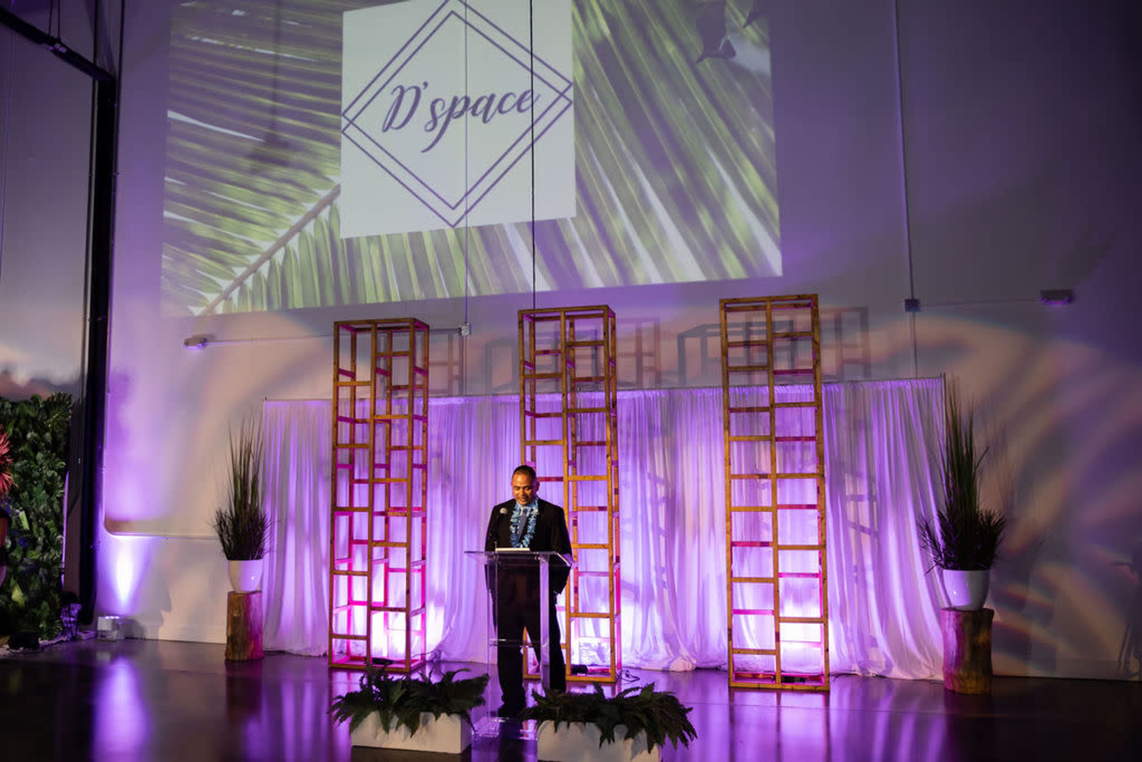 A man stands behind a clear podium on a stage with tropical-themed decorations and lighting while addressing the audience.