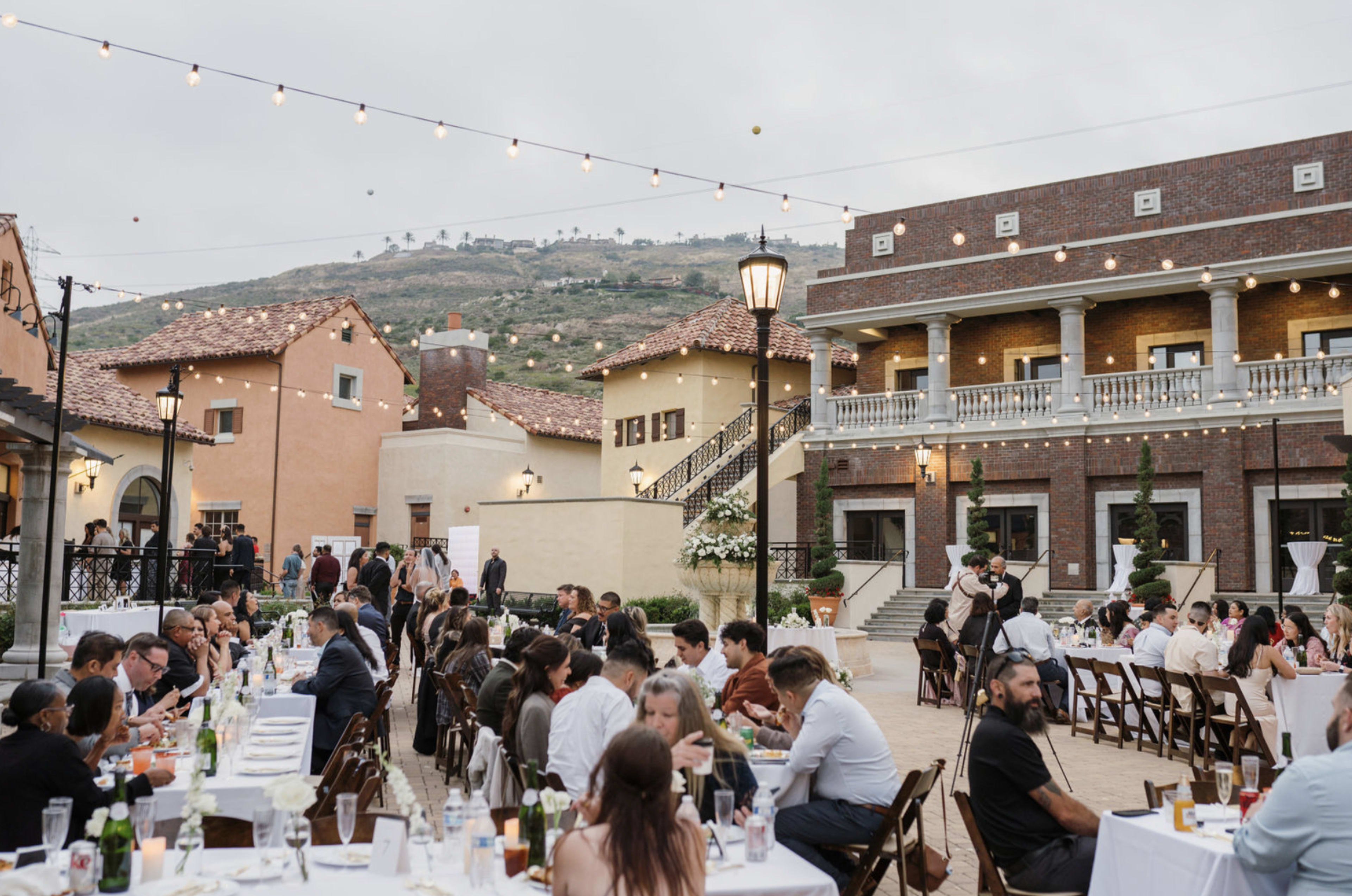 A wedding reception is taking place outdoors in a courtyard surrounded by rustic buildings, with string lights overhead and guests seated at tables.