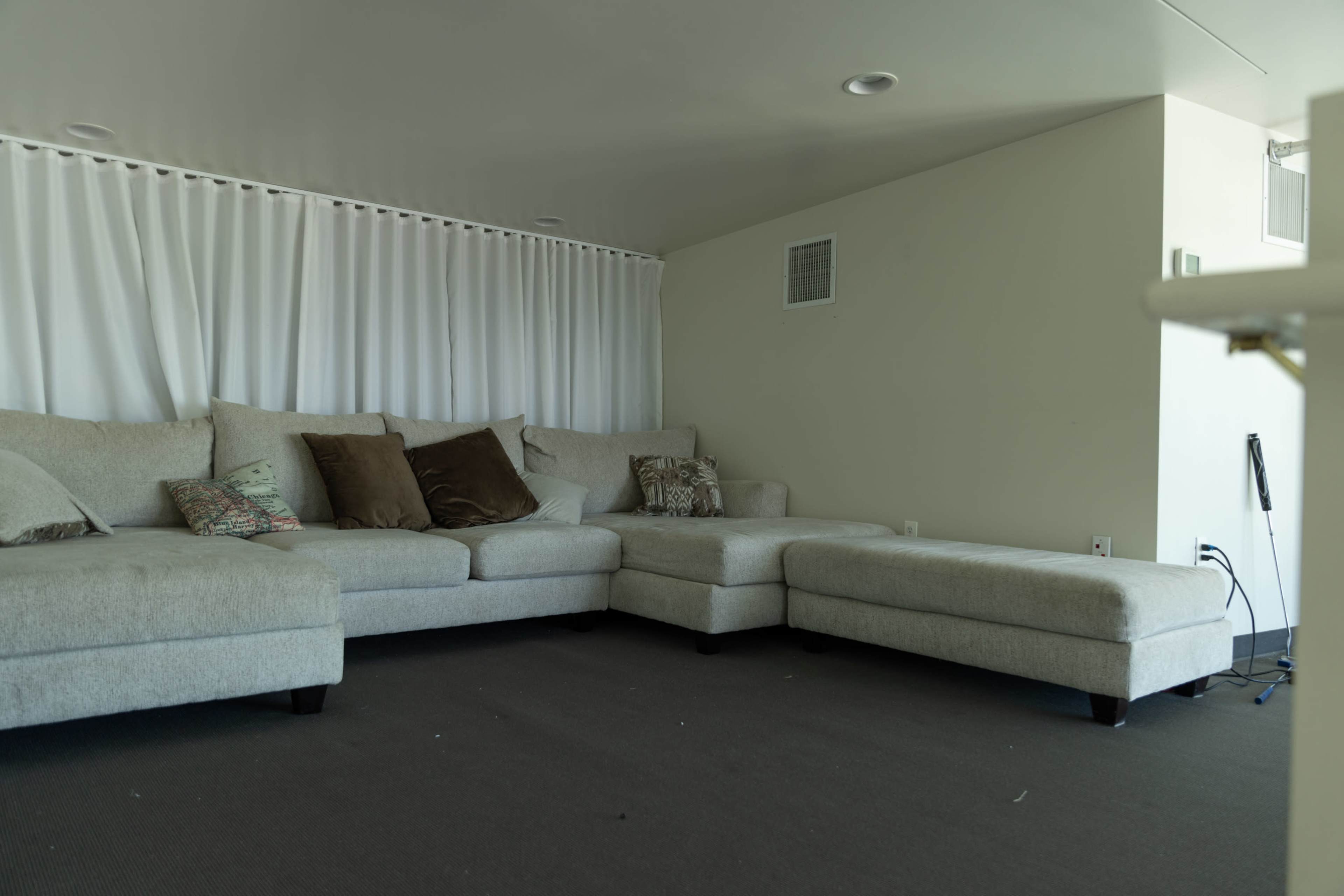 The image shows a light-colored sectional sofa with various pillows, positioned in a sparsely decorated living area.