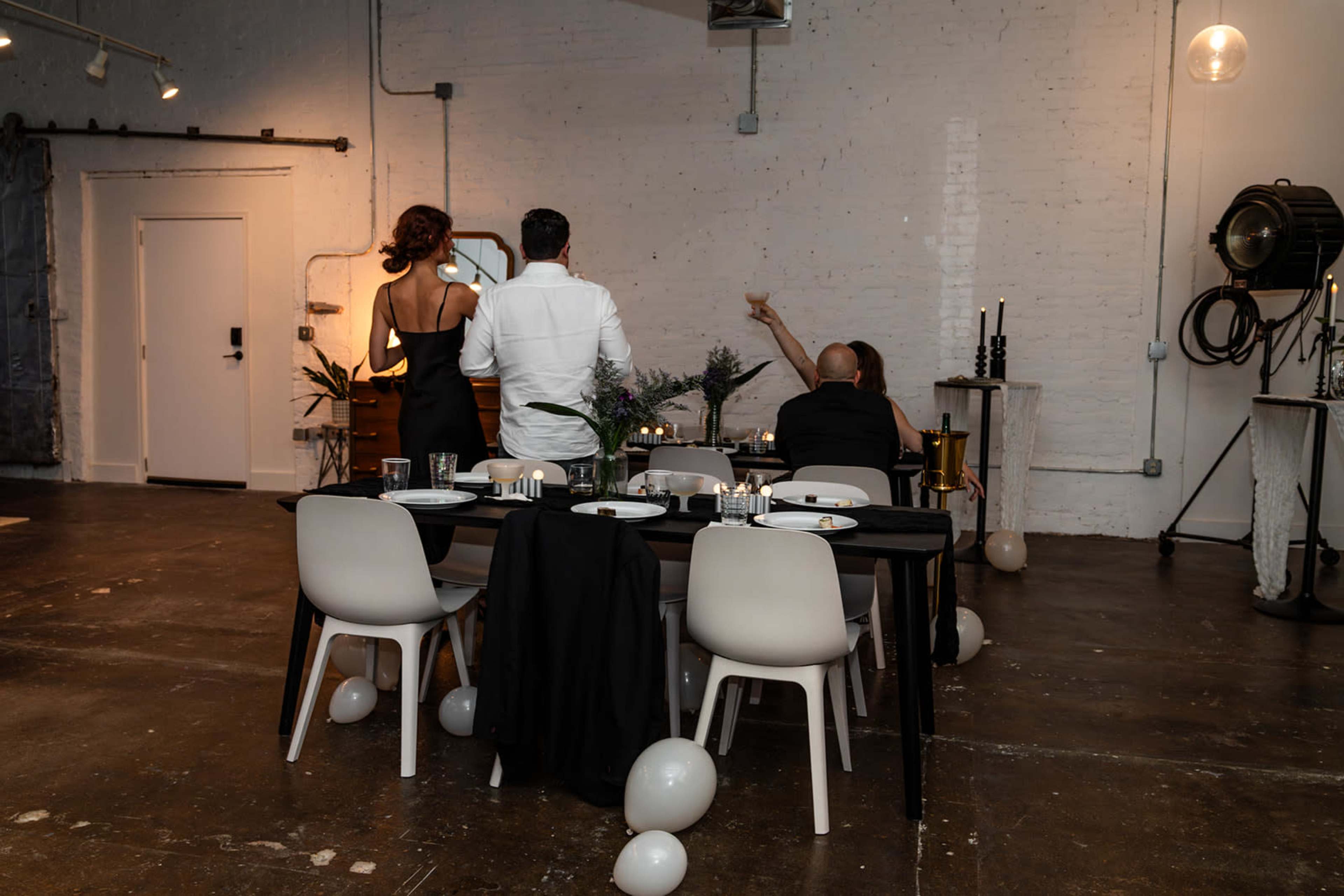 A dining table is set with empty plates and glasses, while three people stand nearby, one gesturing towards a wall in a minimally decorated room.