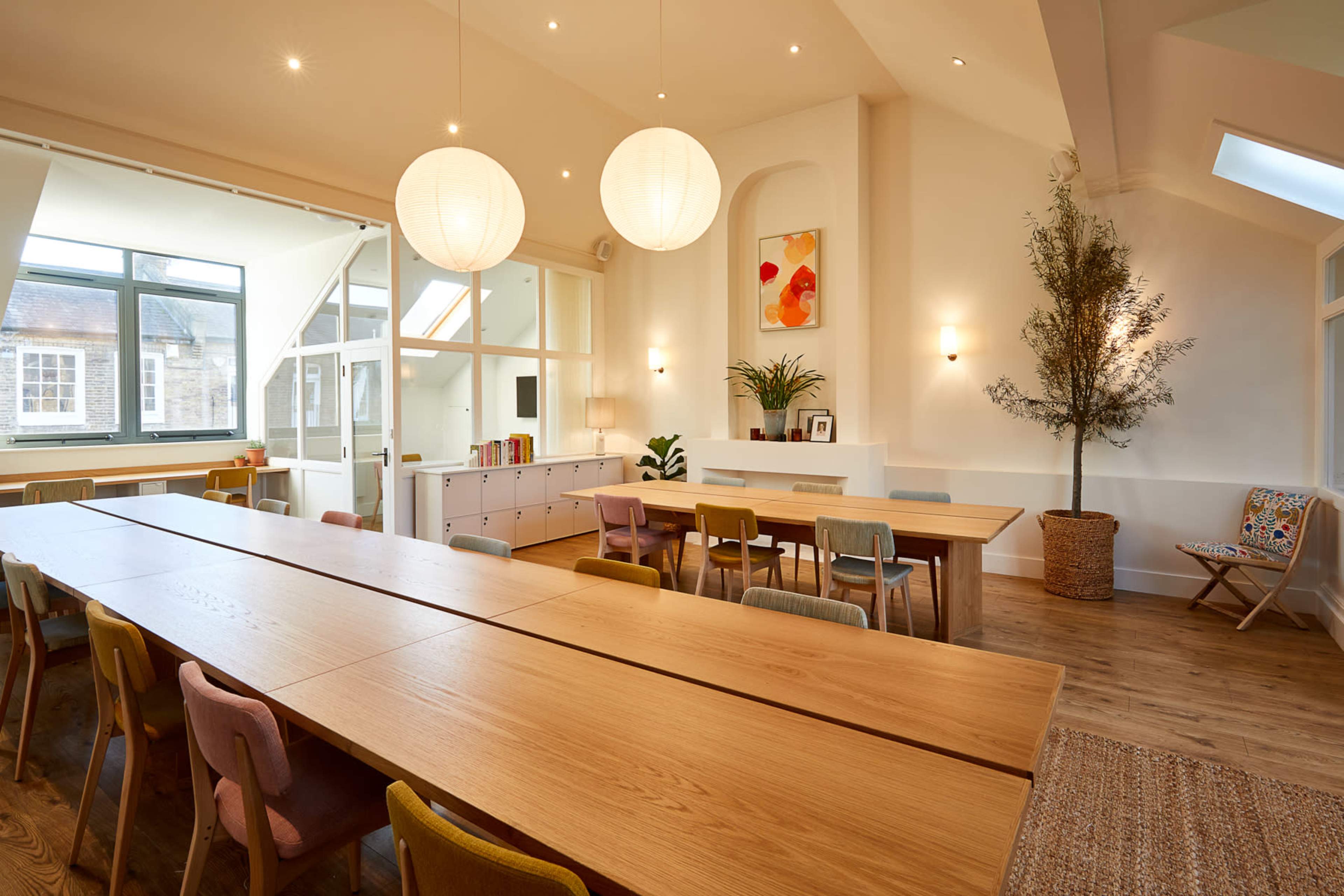 The image shows a spacious, well-lit room featuring a long wooden table with multiple chairs, greenery, and large windows allowing natural light to flood the space.