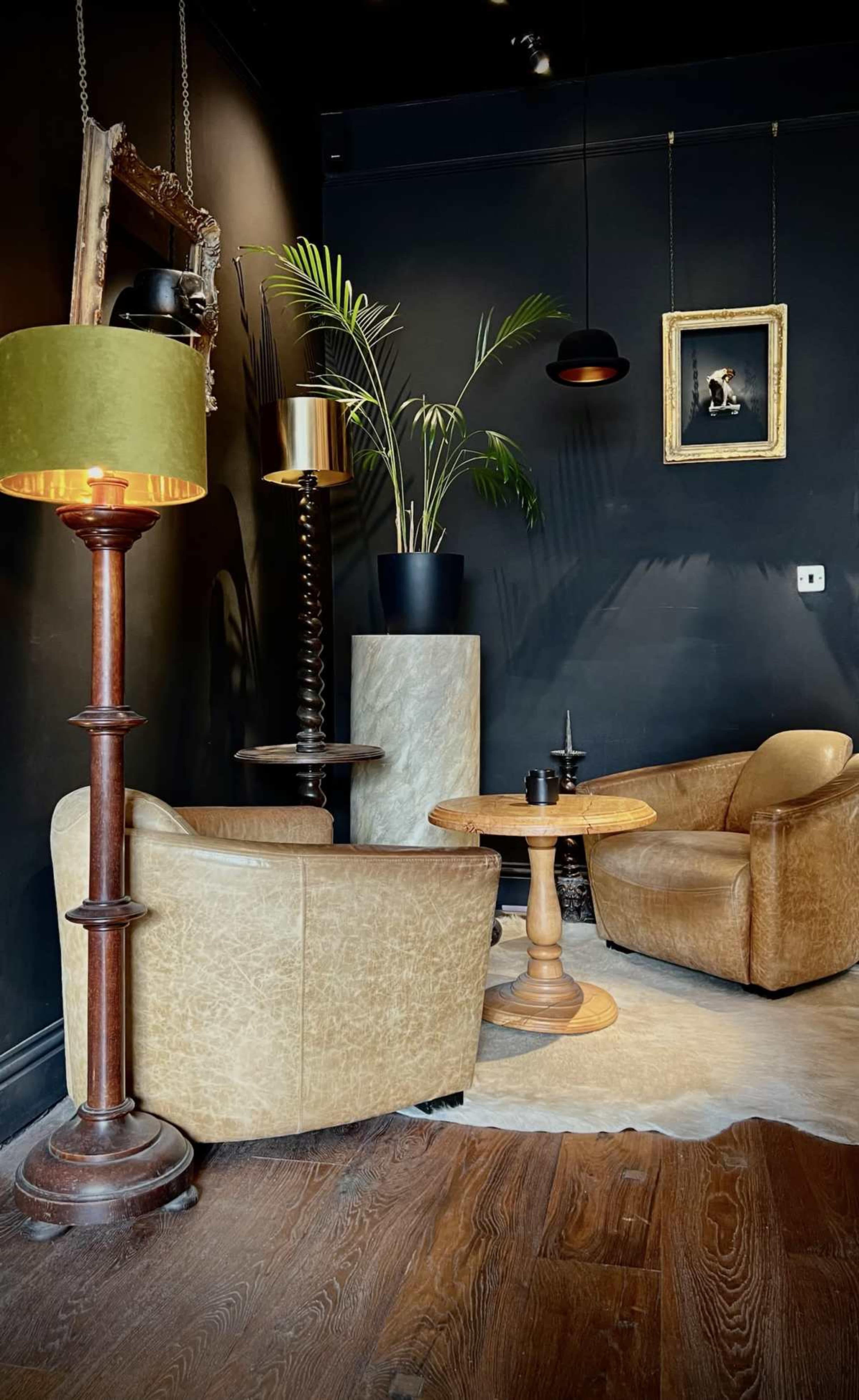 The image features a cozy corner of a room with two vintage leather armchairs, a circular wooden table, a tall lamp, and a potted plant against a dark wall.
