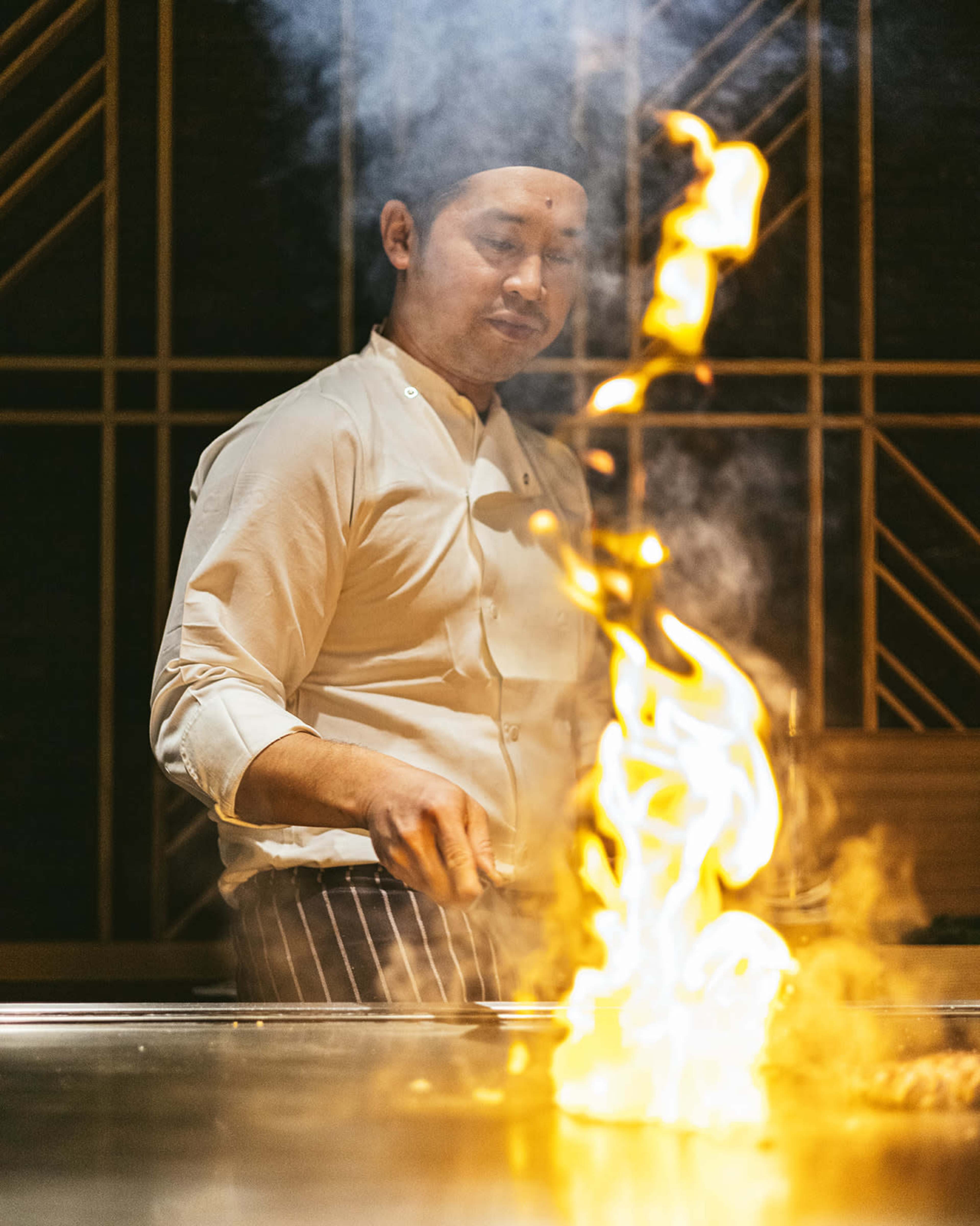 A chef in a white uniform stands in front of a grill, with flames rising as he cooks.